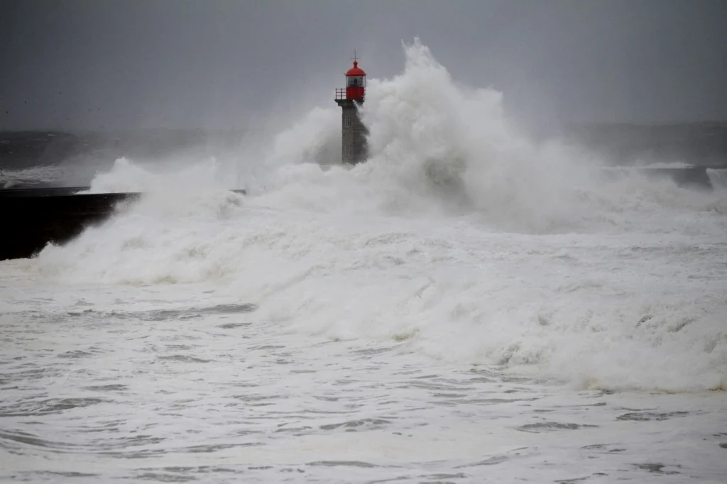 Potężna fala uderza o falochron i latarnię morską z czerwoną kopułą podczas sztormu. Dynamiczny kontrast białych fal z szarością nieba podkreśla siłę żywiołu.