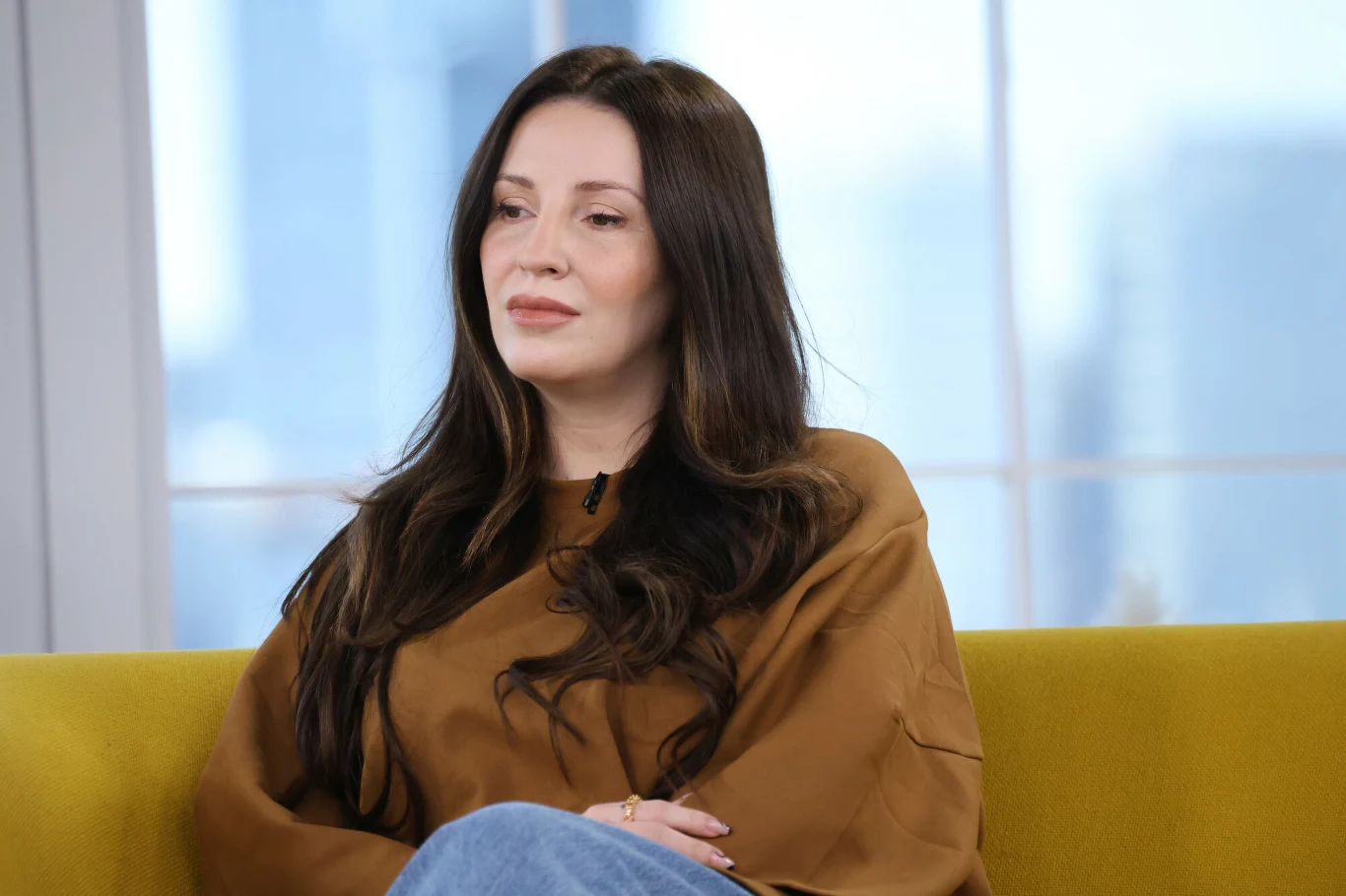 Dominika Serowska Kobieta z długimi ciemnymi włosami siedzi na żółtej kanapie, ubrana w brązową bluzkę i niebieskie spodnie, w tle rozmyte wnętrze z dużymi oknami.