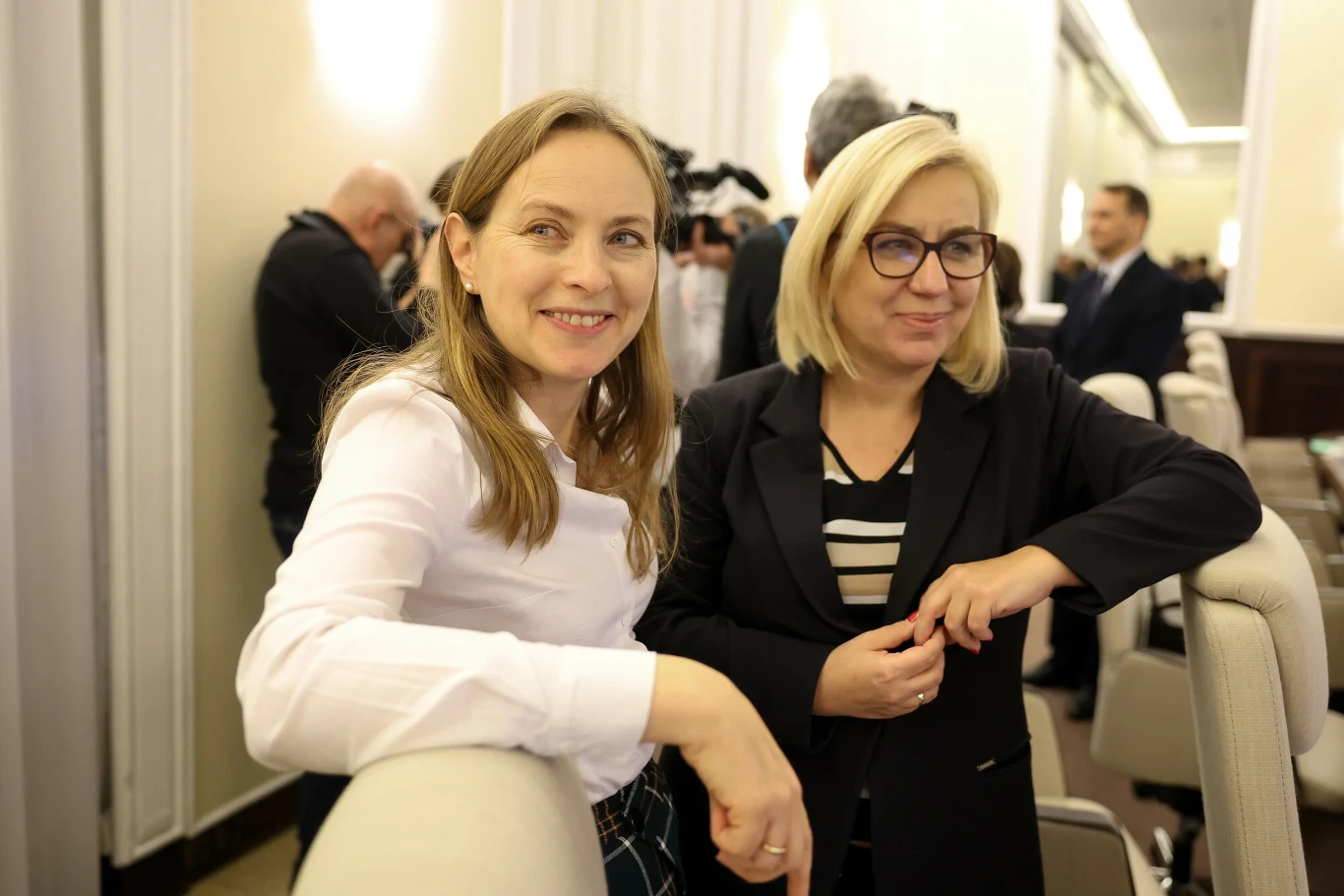 Dwie kobiety stoją obok siebie w eleganckim wnętrzu, jedna uśmiechnięta, druga w okularach, w tle rozmowy i inni uczestnicy spotkania. Atmosfera formalna, wnętrze jasne i nowoczesne.