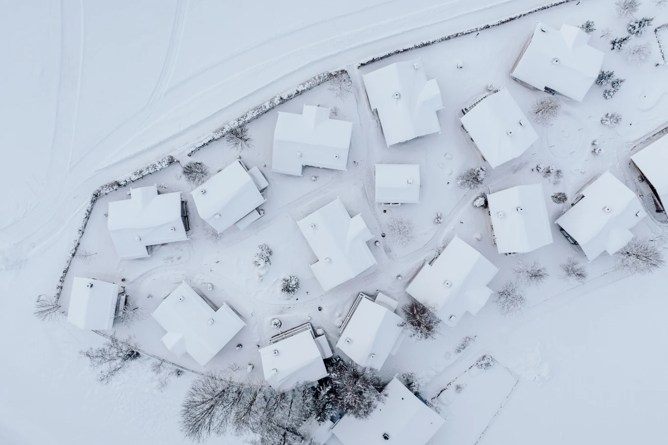 Osiedle domów jednorodzinnych pokrytych grubą warstwą śniegu, sfotografowane z lotu ptaka. Ulice i dachy całkowicie przykryte bielą, otoczone zamarzniętymi drzewami i ogrodzeniami.