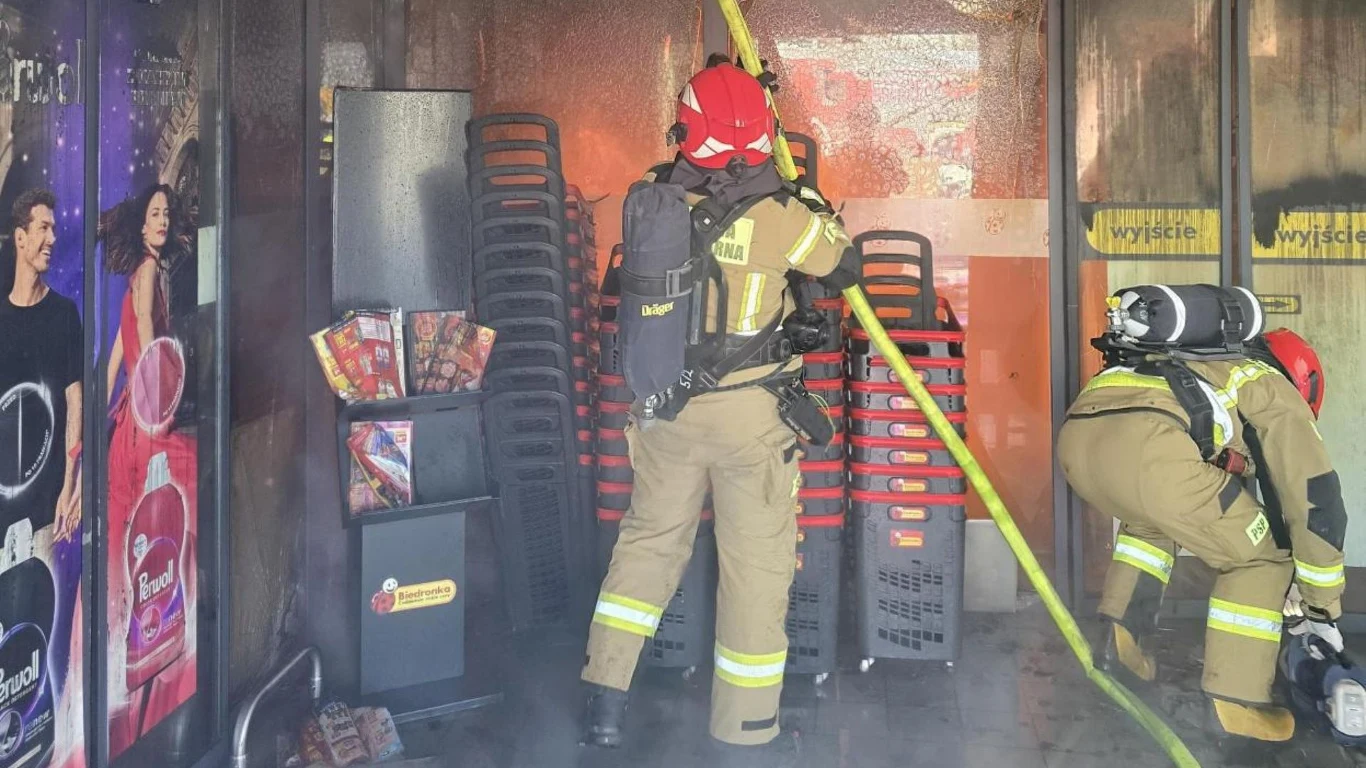Straży walczą z pożarem w Polkowicach Dwóch strażaków w pełnym umundurowaniu oraz maskach ochronnych gasi pożar wewnątrz sklepu, jeden z nich trzyma wąż strażacki skierowany w stronę źródła ognia, wokół widoczne zadymienie oraz plastikowe koszyki na zakupy.