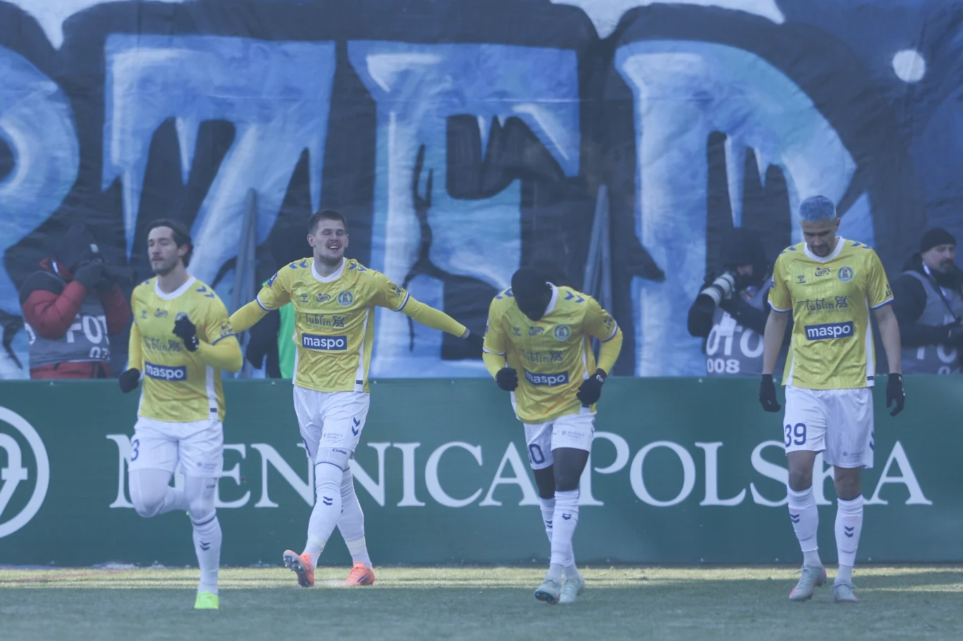 Czterech piłkarzy w żółtych koszulkach i białych spodenkach biegnie wzdłuż murawy na tle wielkiego, niebieskiego graffiti oraz baneru reklamowego z napisem Mennica Polska. Rozgrywane wydarzenie odbywa się na odkrytym stadionie w chłodnej aurze.