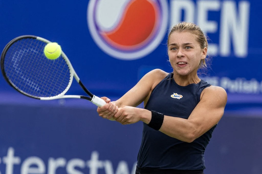 Maja Chwalińska zagra w Wimbledonie. Tenisistka podczas meczu koncentrująca się na odbiciu żółtej piłki rakietą. W tle niebieski baner turnieju tenisowego.