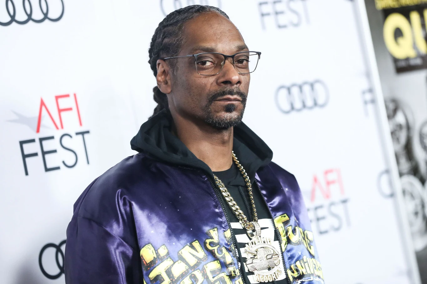 Snoop Dogg Mężczyzna w okularach i marynarce z naszyjnikami stoi na tle ścianki z logo AFI FEST oraz logo Audi, tło jasne, motyw wydarzenia festiwalowego.