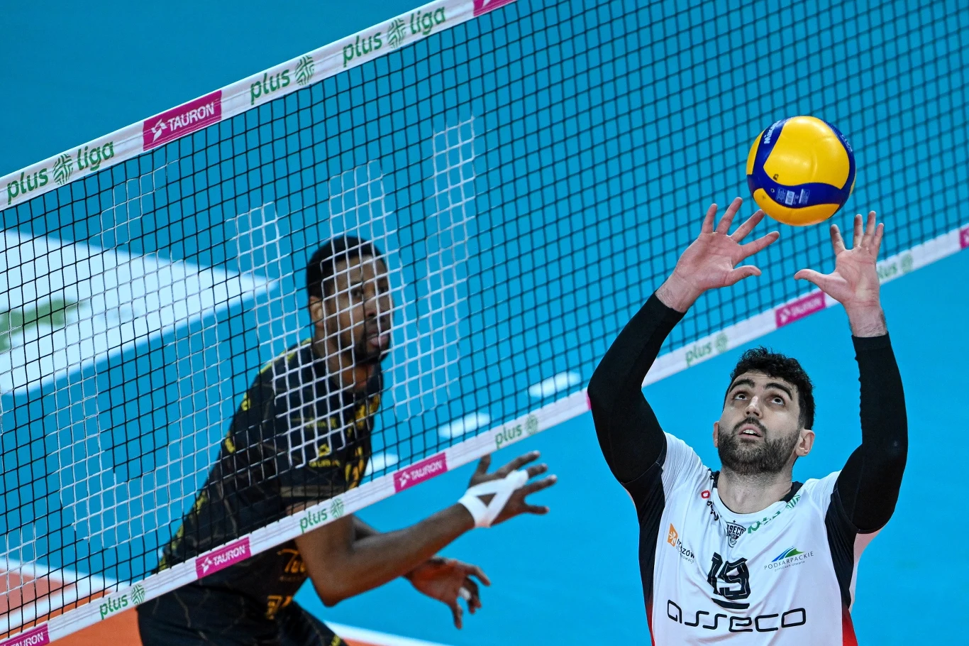 Zawodnik drużyny Asseco Resovia podnosi piłkę do ataku podczas meczu siatkówki, drugi zawodnik w tle przygotowuje się do akcji przy siatce.