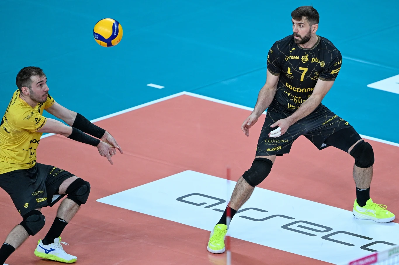 Dwóch siatkarzy w żółto-czarnych strojach podczas akcji na boisku, jeden z graczy przygotowuje się do przyjęcia piłki, na tle niebieskiego parkietu i logotypów na podłodze.