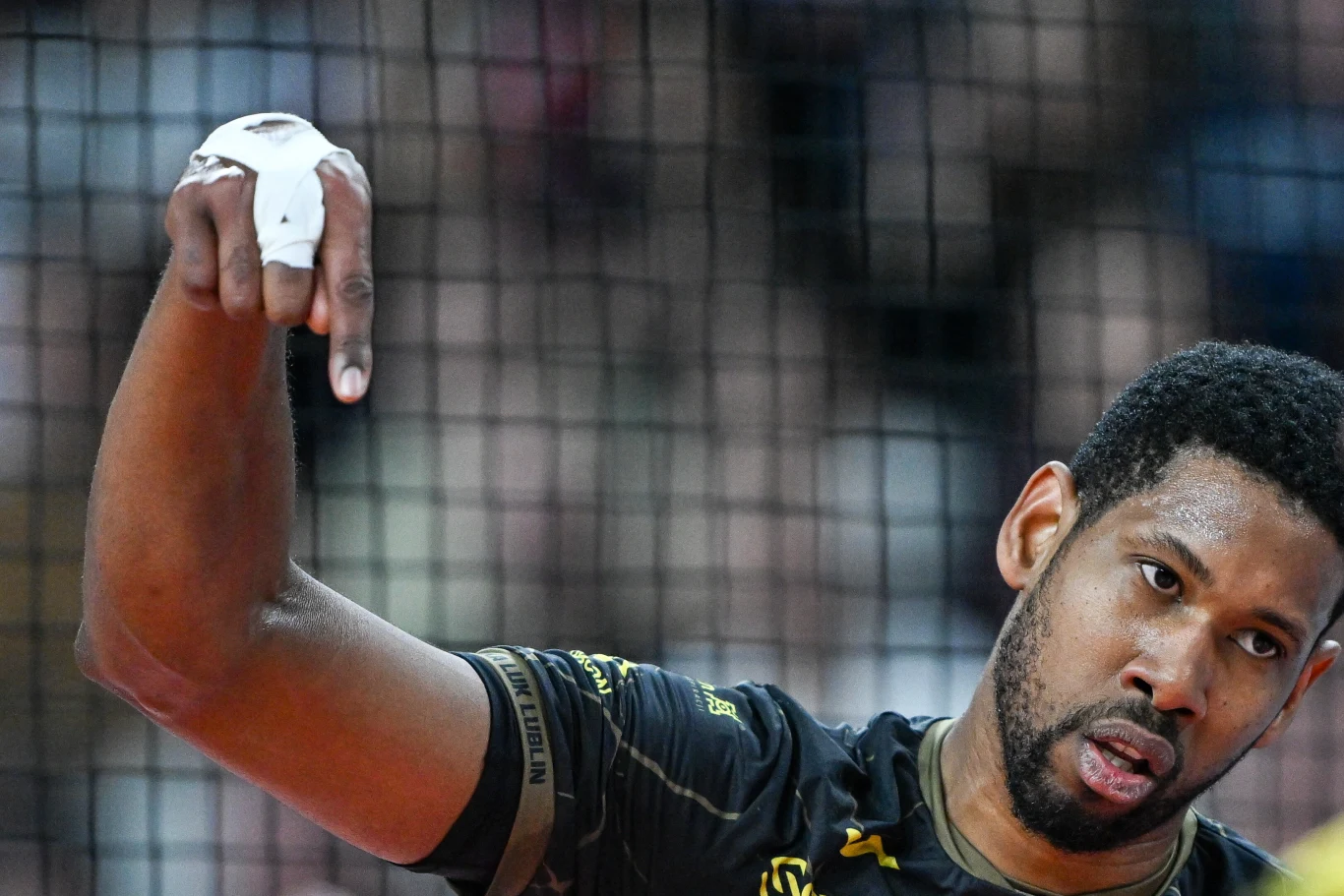 Zbliżenie na sportowca w czarnej koszulce z owiniętym bandażem na palcach, wykonującego gest dłonią na tle siatki do siatkówki.