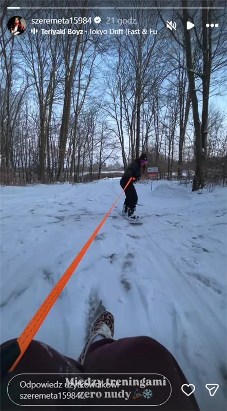 Osoba ciągnie drugą osobę siedzącą na sankach po zaśnieżonej leśnej ścieżce, wokół widoczne wysokie, bezlistne drzewa.
