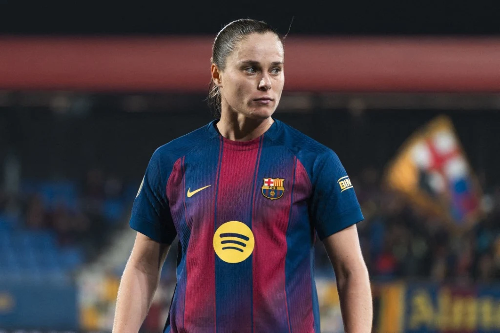 Piłkarka w stroju FC Barcelona z logo sponsora na piersi, patrząca w bok na tle stadionu z flagą klubową w tle.