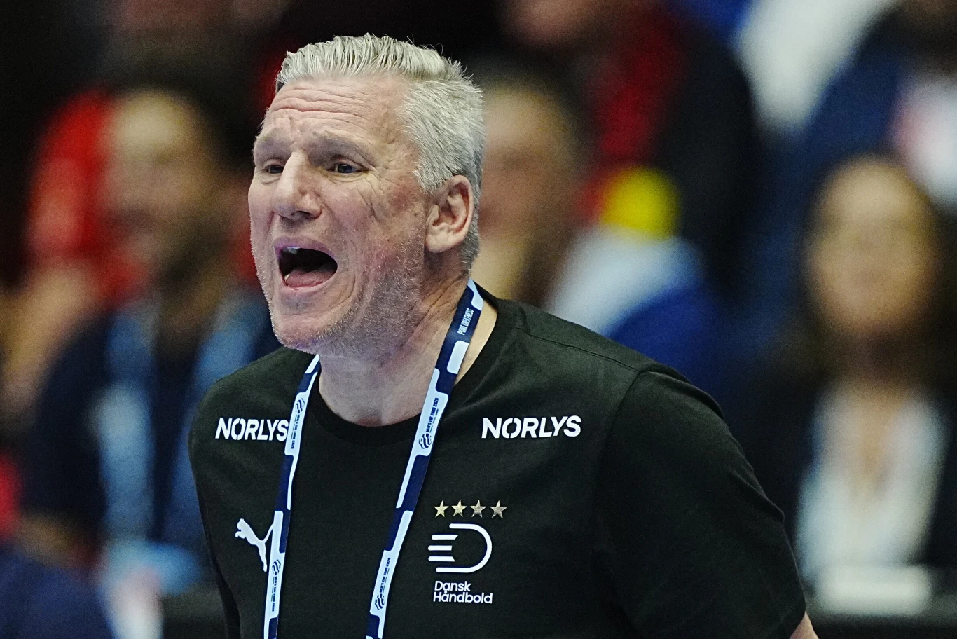 Mężczyzna z siwymi włosami w czarnej koszulce z logo i napisem Damsk Håndbold stoi z otwartymi ustami, prawdopodobnie w trakcie okrzyku lub wydawania poleceń, w tle rozmazana publiczność.