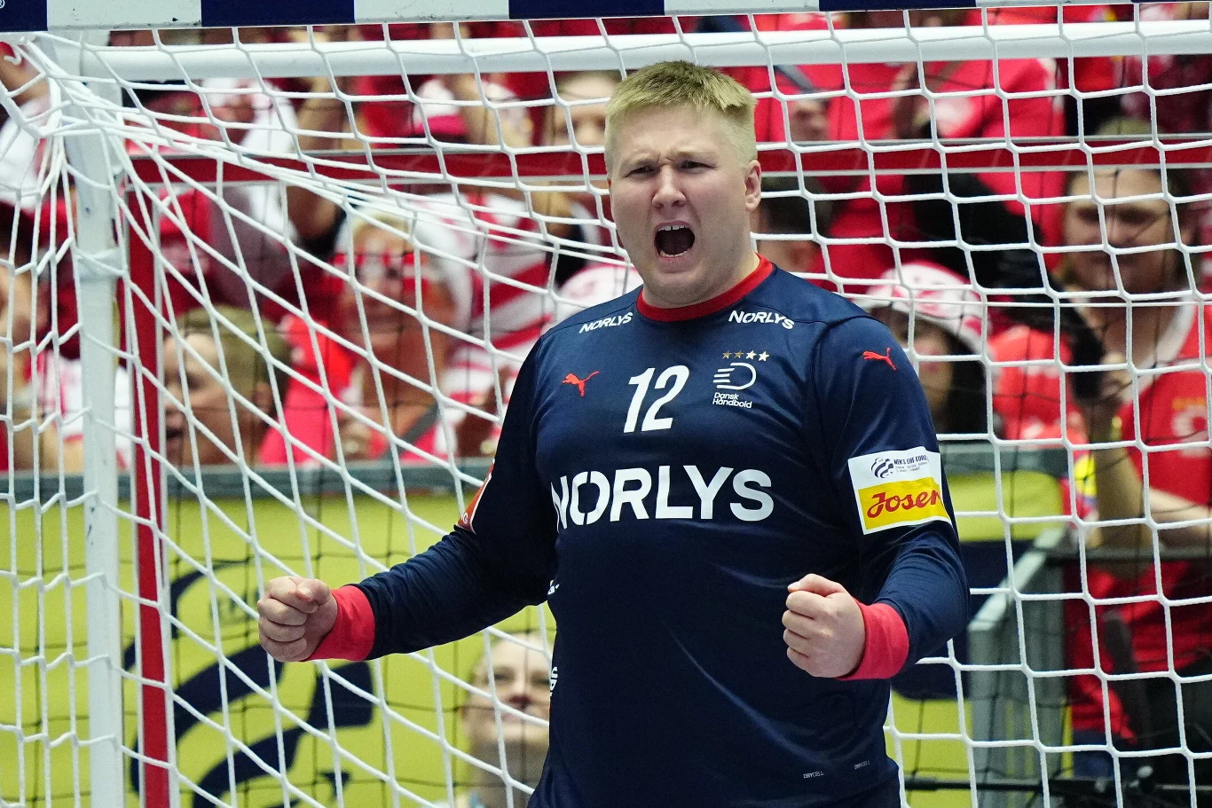 Bramkarz drużyny piłki ręcznej w granatowym stroju z numerem 12, unosi obie pięści w geście triumfu. W tle widoczne są trybuny z kibicami u ubraniach w barwach narodowych.