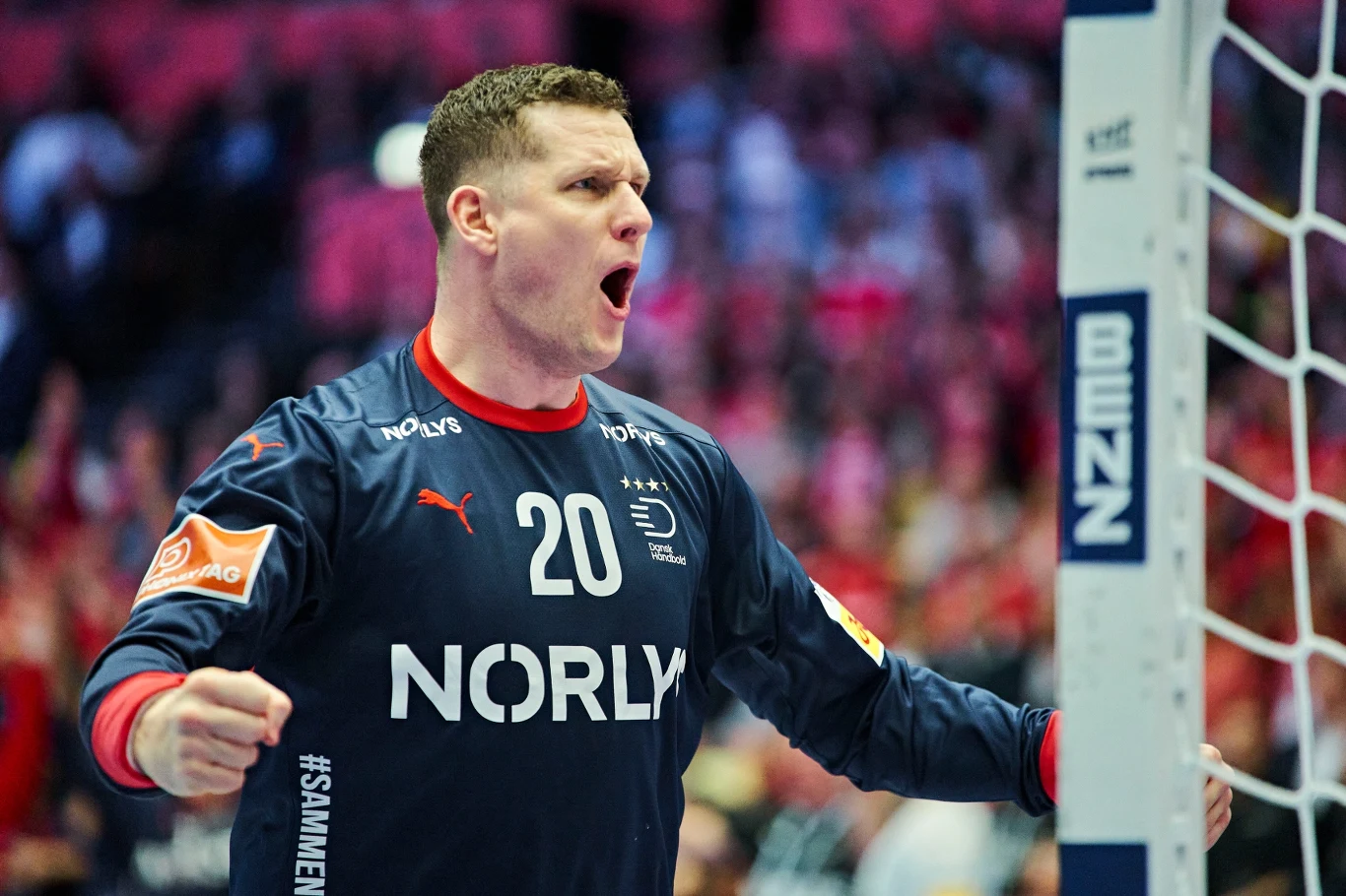 Zawodnik piłki ręcznej w stroju bramkarza z numerem 20, z uniesionymi pięściami i wyrażającym silne emocje wyrazem twarzy. W tle rozmyte postacie kibiców i fragment bramki.