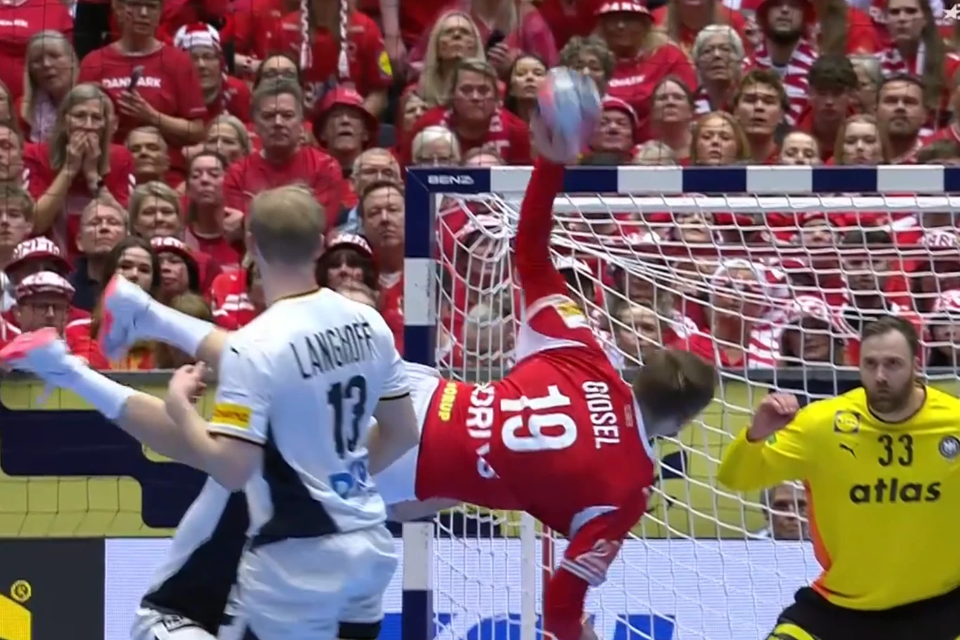 Zawodnik w czerwonym stroju wykonuje spektakularny rzut podczas meczu piłki ręcznej, próbując zdobyć bramkę pomimo obecności obrońcy w białej koszulce oraz bramkarza bacznie obserwującego sytuację na tle pełnych trybun kibiców.