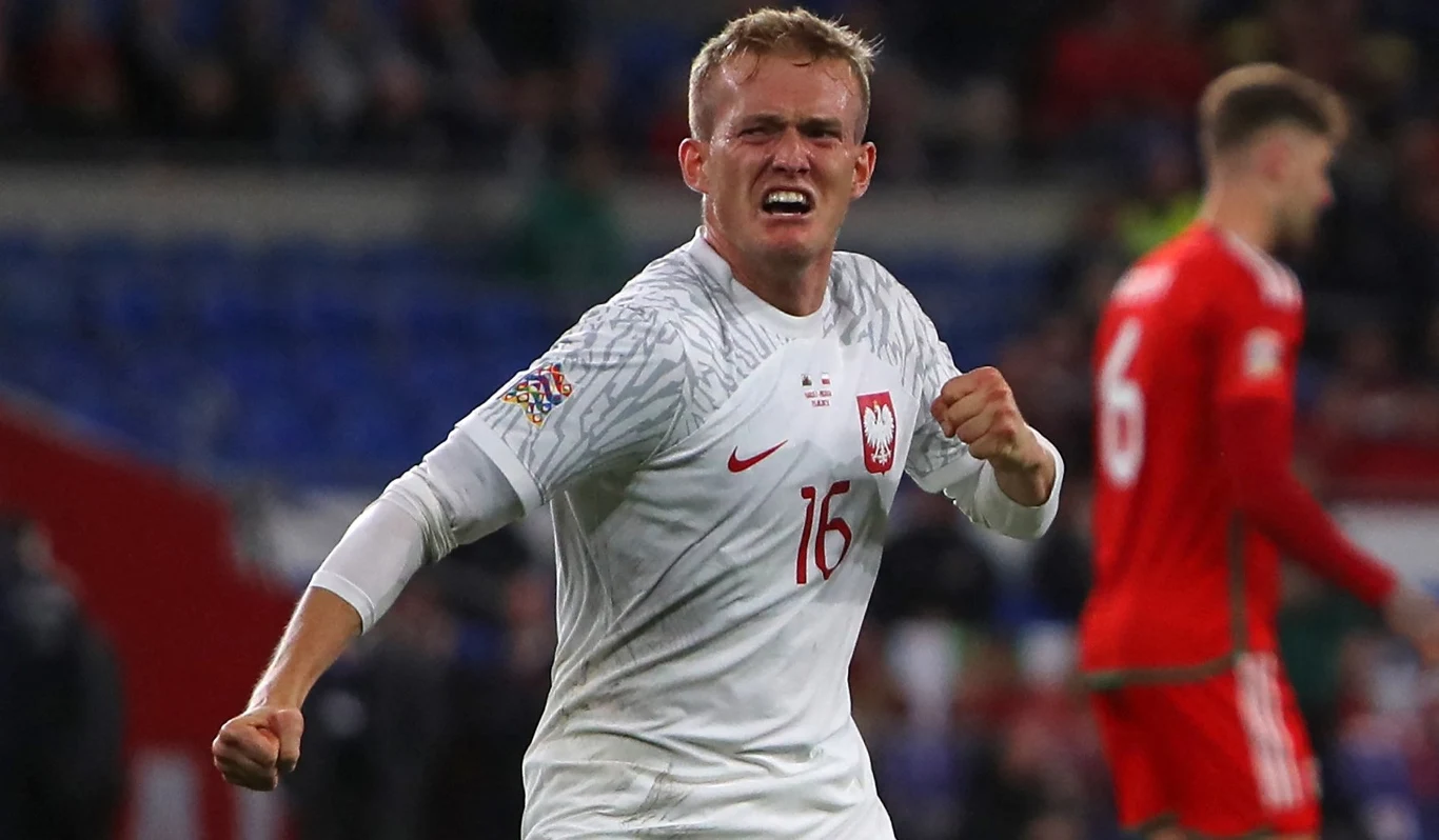 Polski piłkarz w białej koszulce z numerem 16 świętuje sukces na boisku, zaciśnięta pięść i wyraz twarzy pokazujący silne emocje. W tle znajduje się zawodnik w czerwonym stroju.