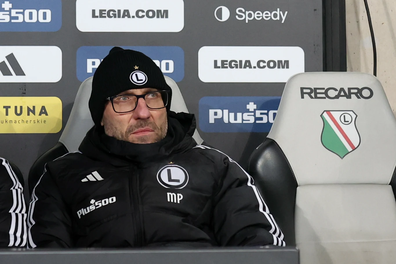 Trener drużyny piłkarskiej Legia Warszawa siedzący na ławce rezerwowych, ubrany w klubową kurtkę i czapkę z logo zespołu, za nim plansze ze sponsorami.