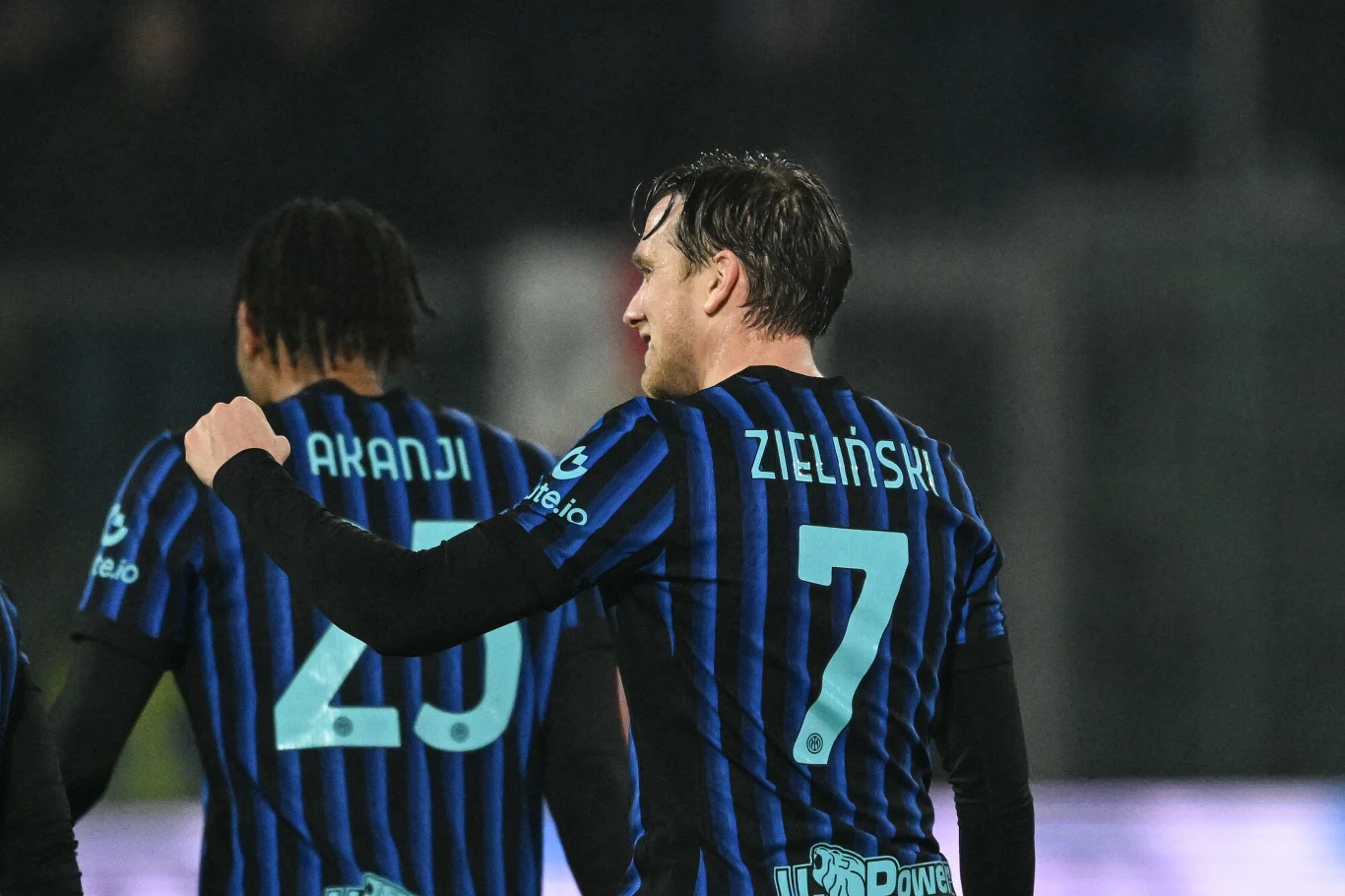 Dwóch piłkarzy ubranych w czarno-niebieskie stroje drużyny piłkarskiej, z widocznymi nazwiskami Akanji oraz Zieliński i numerami na plecach, podczas meczu na stadionie.