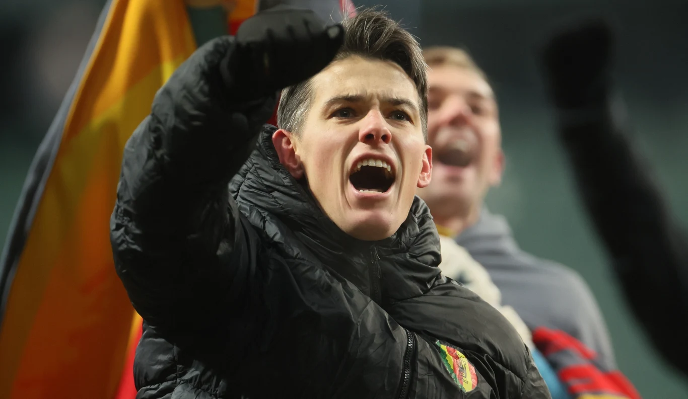 Młody człowiek ubrany w ciemną kurtkę z podniesioną pięścią wydaje się głośno wyrażać emocje, w tle widoczny rozmyty tłum oraz kolorowa flaga.