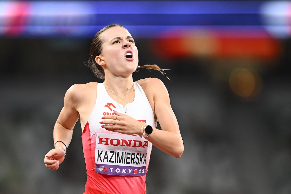 Lekkoatletka w stroju sportowym podczas biegu, widoczna determinacja na twarzy oraz napięcie mięśni. Na koszulce nazwisko zawodniczki oraz logotypy sponsorów.