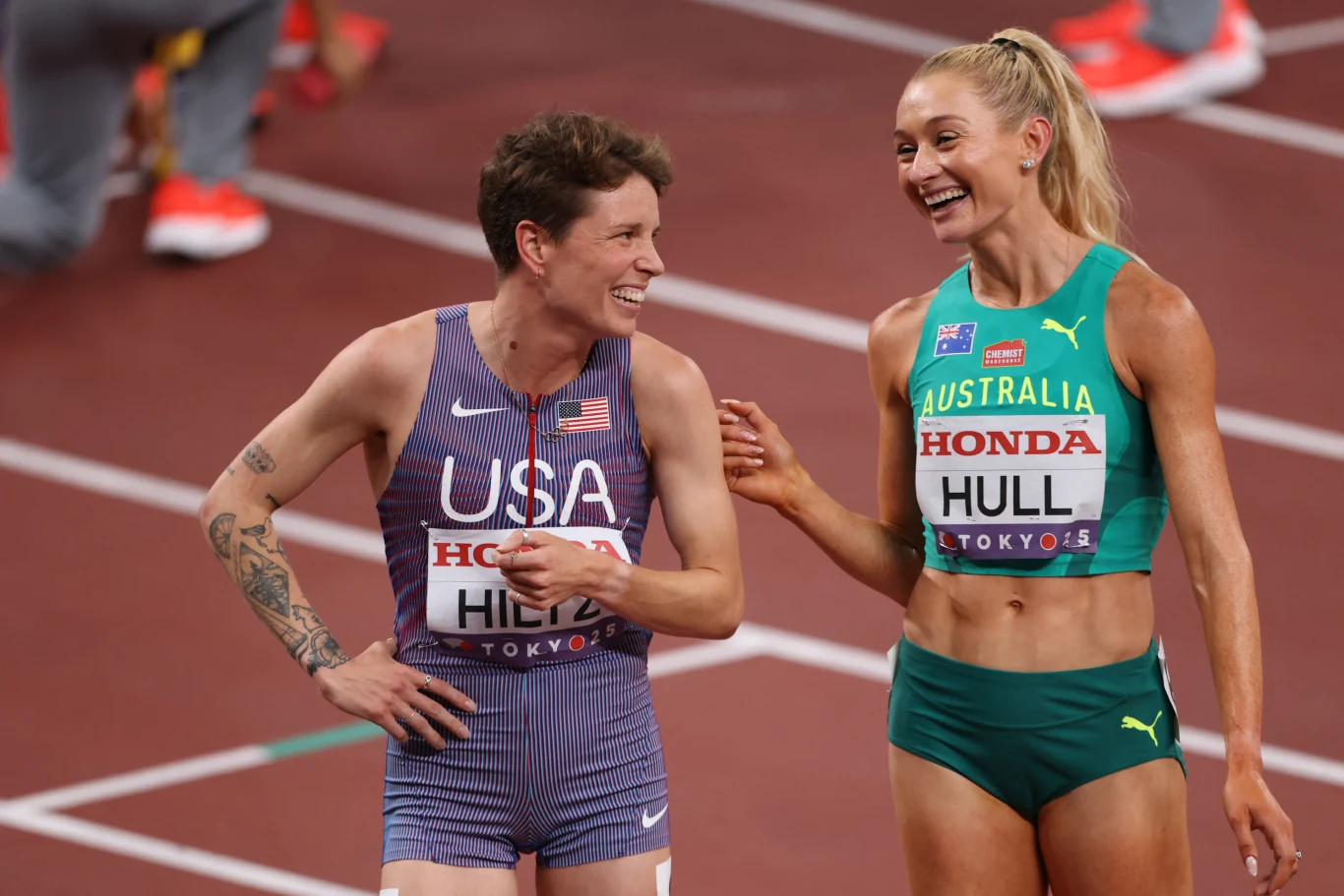 Dwie lekkoatletki w sportowych strojach reprezentujących USA i Australię rozmawiają i śmieją się na bieżni stadionu lekkoatletycznego po zakończonym biegu.
