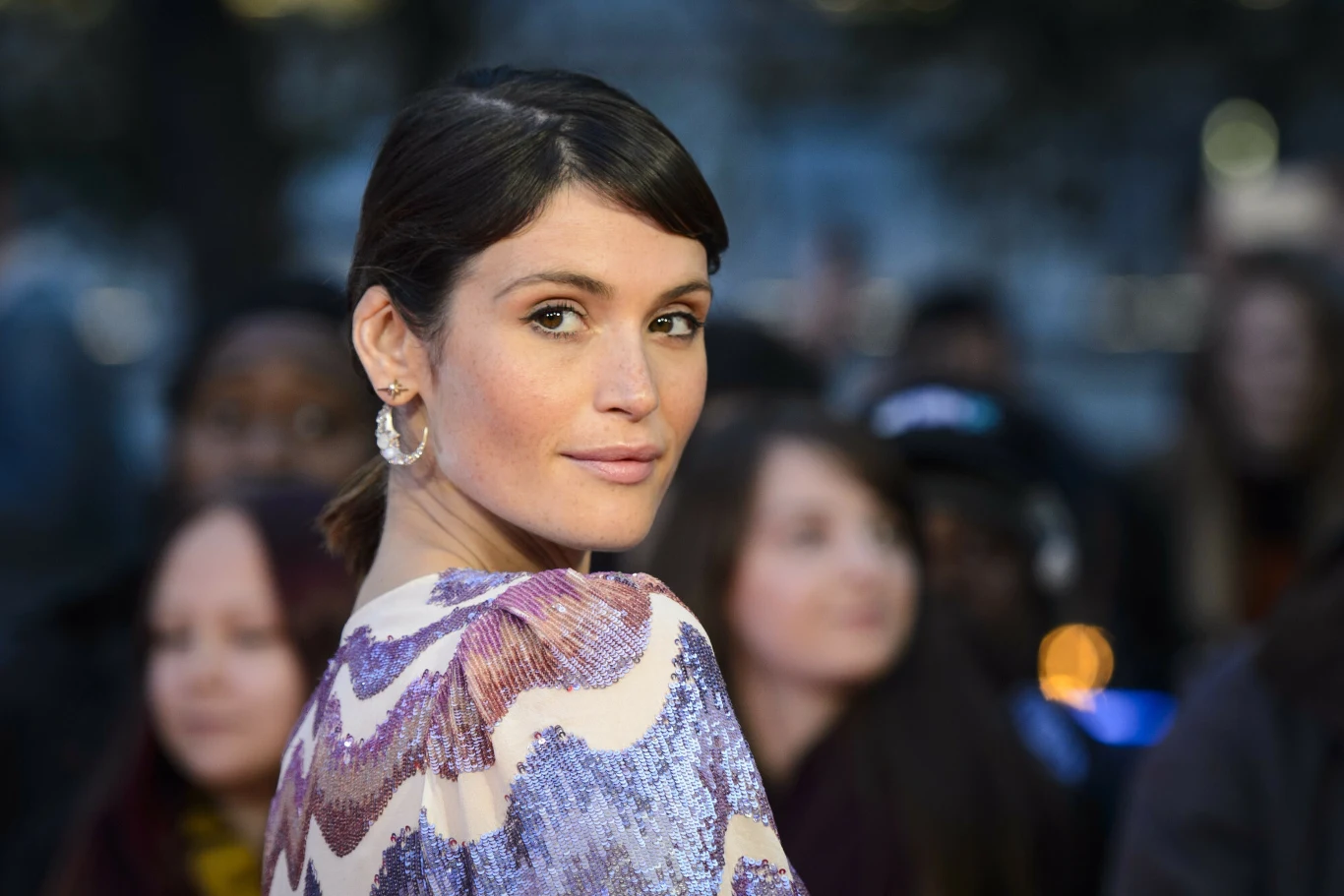 Kobieta z ciemnymi włosami upiętymi do tyłu, uśmiecha się delikatnie i patrzy przez ramię w stronę aparatu. Ubrana w błyszczącą bluzkę z fioletowo-białym wzorem oraz srebrne kolczyki. W tle rozmyta grupa osób oraz światła miasta.