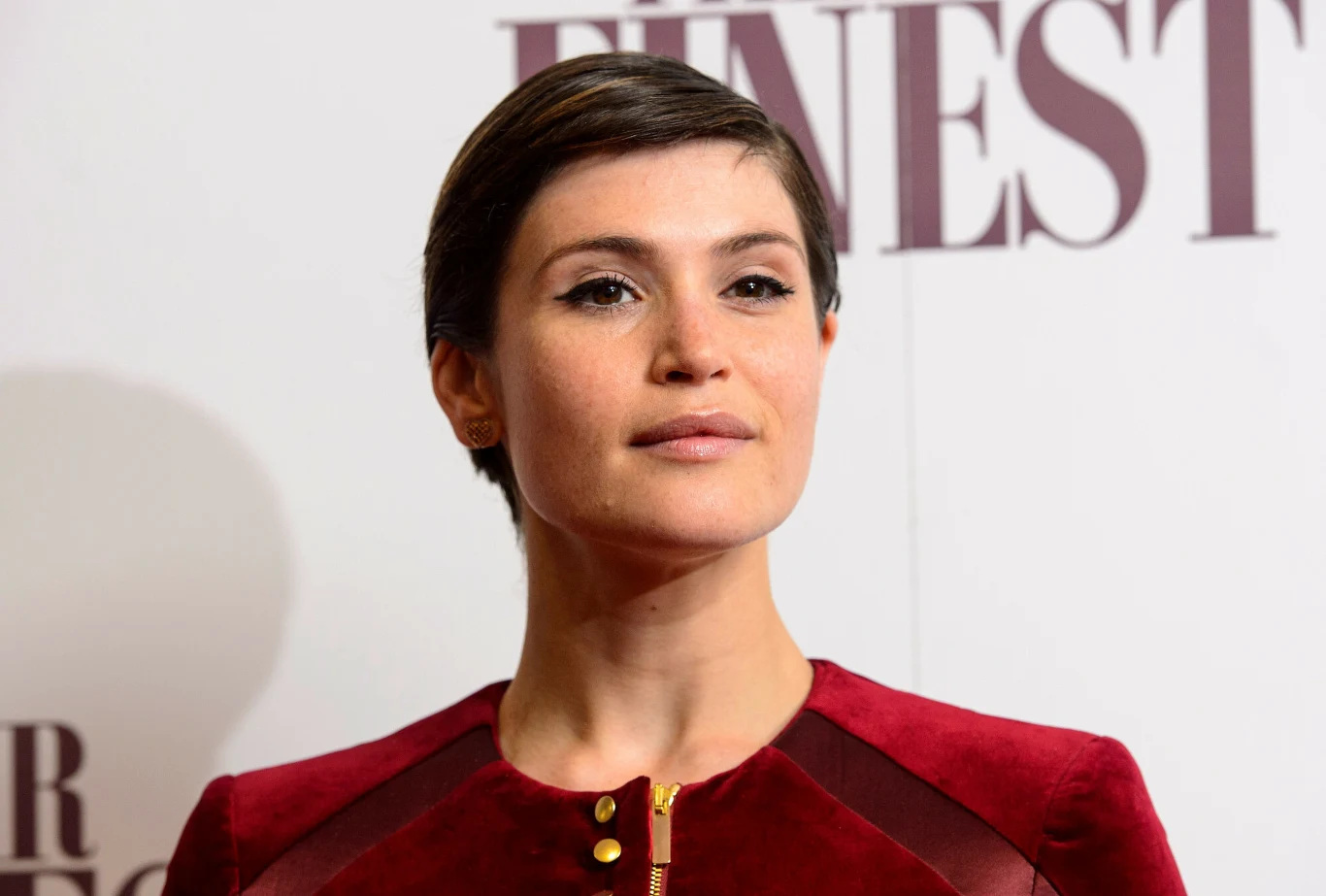 Kobieta o krótkich, ciemnych włosach ubrana w bordowy elegancki strój stoi na tle białej ścianki z napisem. Wygląda pewnie i spokojnie, patrzy lekko w bok.