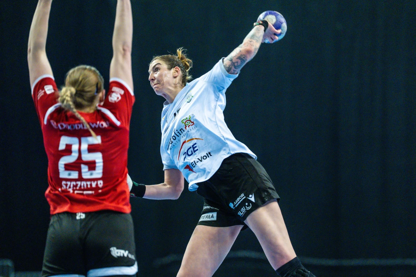 Kadr z meczu Orlen Superligi Kobiet Dwie zawodniczki piłki ręcznej w trakcie dynamicznej akcji meczowej, jedna w czerwonym stroju podnosi ręce do góry blokując, druga w białej koszulce szykuje się do oddania rzutu piłką.