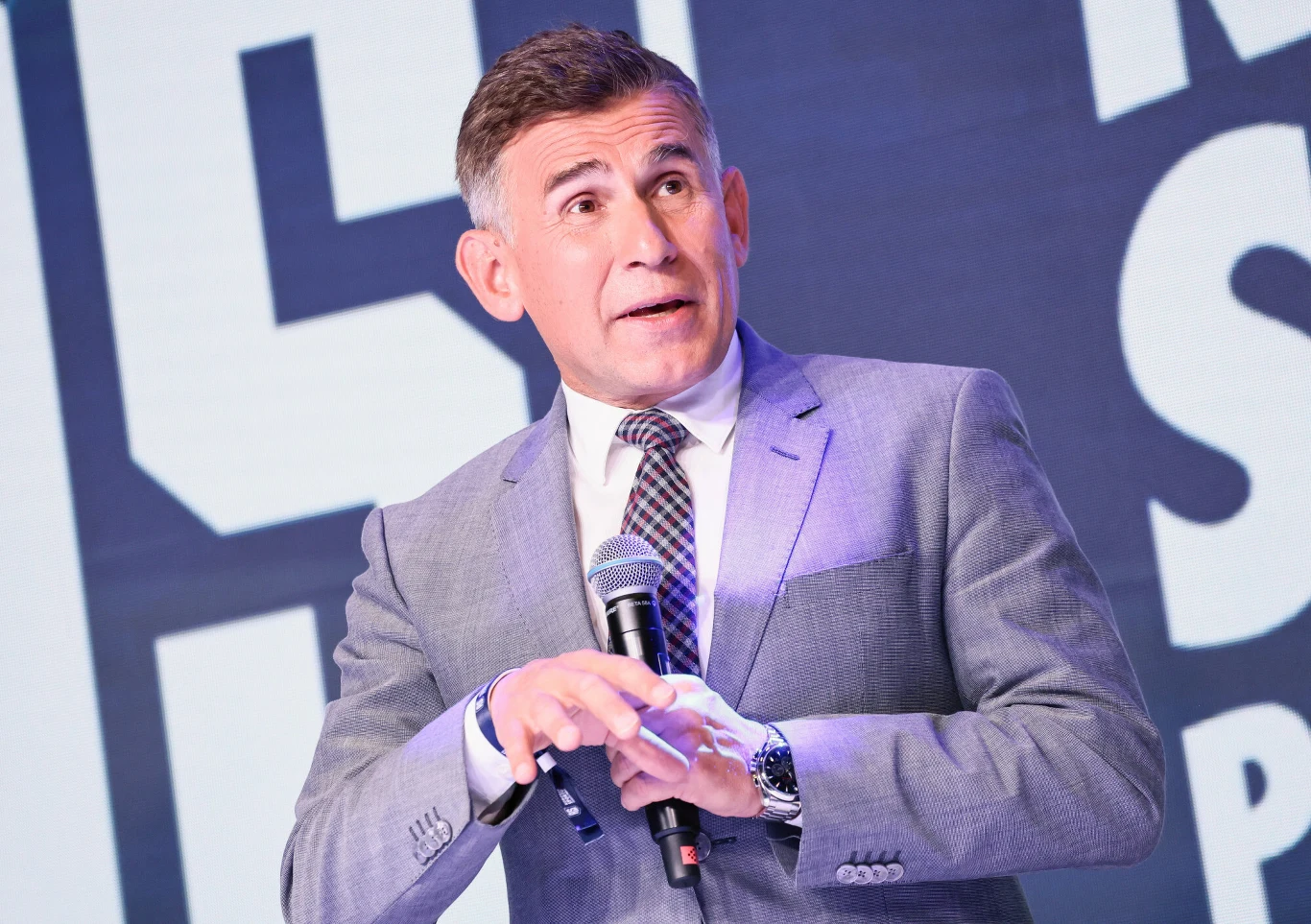 Mężczyzna w garniturze trzyma mikrofon i przemawia na tle dużego, nowoczesnego napisu. Wyraz twarzy sugeruje zaangażowanie i skupienie, a formalny strój podkreśla profesjonalizm sytuacji.