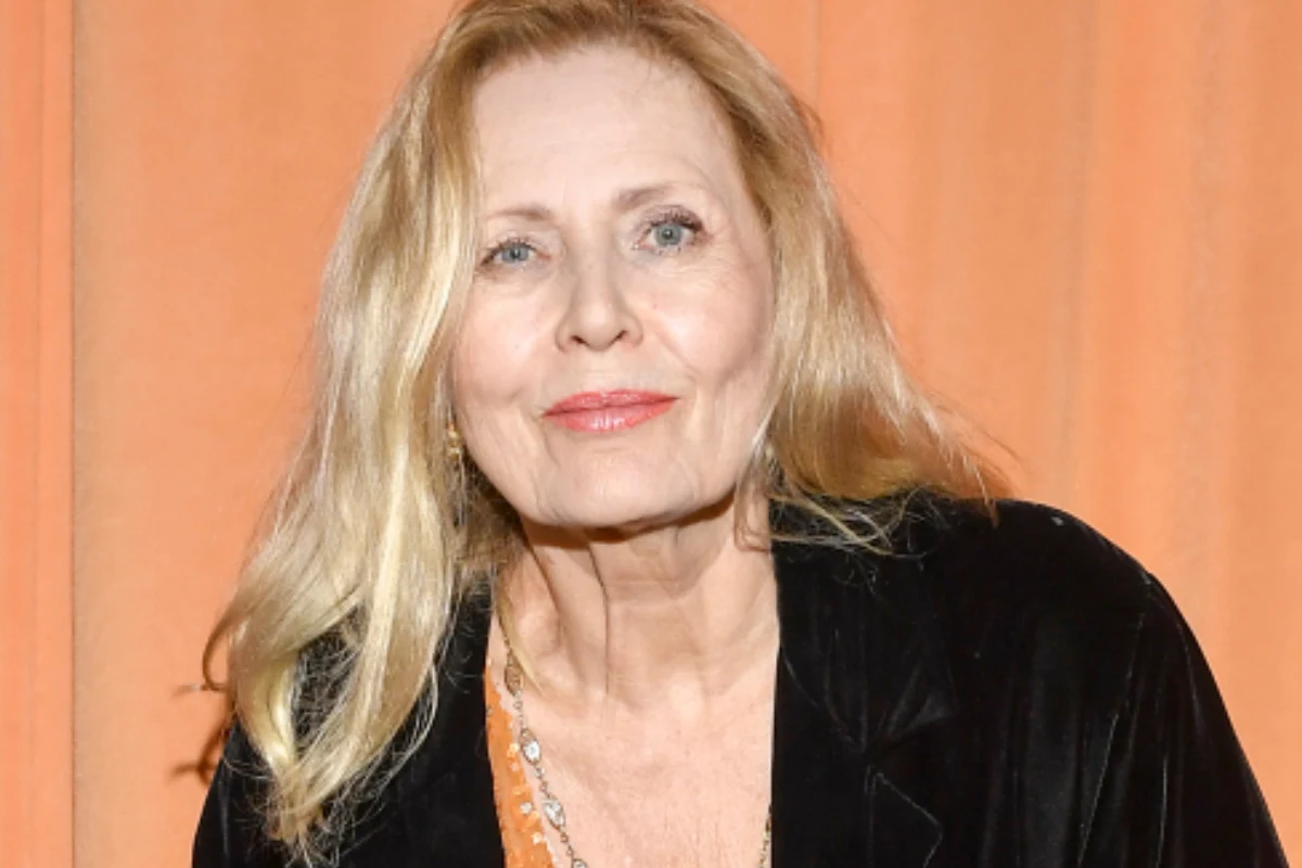 Grażyna Szapołowska Kobieta o blond włosach i jasnej karnacji, ubrana w czarną marynarkę oraz pomarańczową bluzkę, stoi na tle pomarańczowej kurtyny, z delikatnym uśmiechem na twarzy.