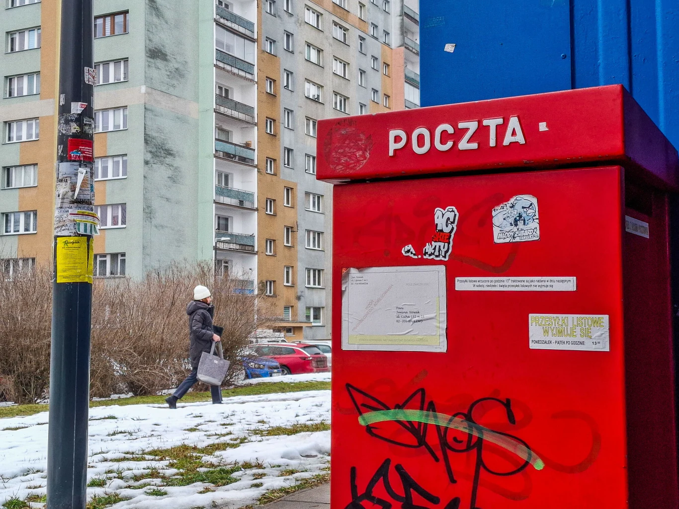 e-Doręczenia uratowały Pocztę Polską? Wiceminister zabrał głos (zdj. ilustracyjne) Czerwona skrzynka pocztowa z napisem „POCZTA” ustawiona przy chodniku na tle bloków mieszkalnych. Skrzynka jest pokryta naklejkami i graffiti. W tle osoba idzie po chodniku, widoczne są resztki śniegu, trawa oraz kilka zaparkowanych samochodów.