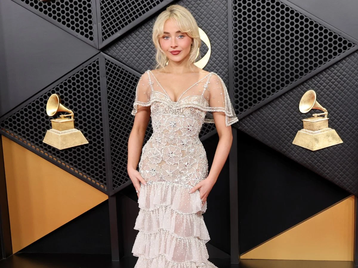 Młoda kobieta w eleganckiej, połyskującej sukni z przezroczystą górą pozuje na tle dekoracji z motywami złotych gramofonów, będących symbolem nagrody muzycznej Grammy.