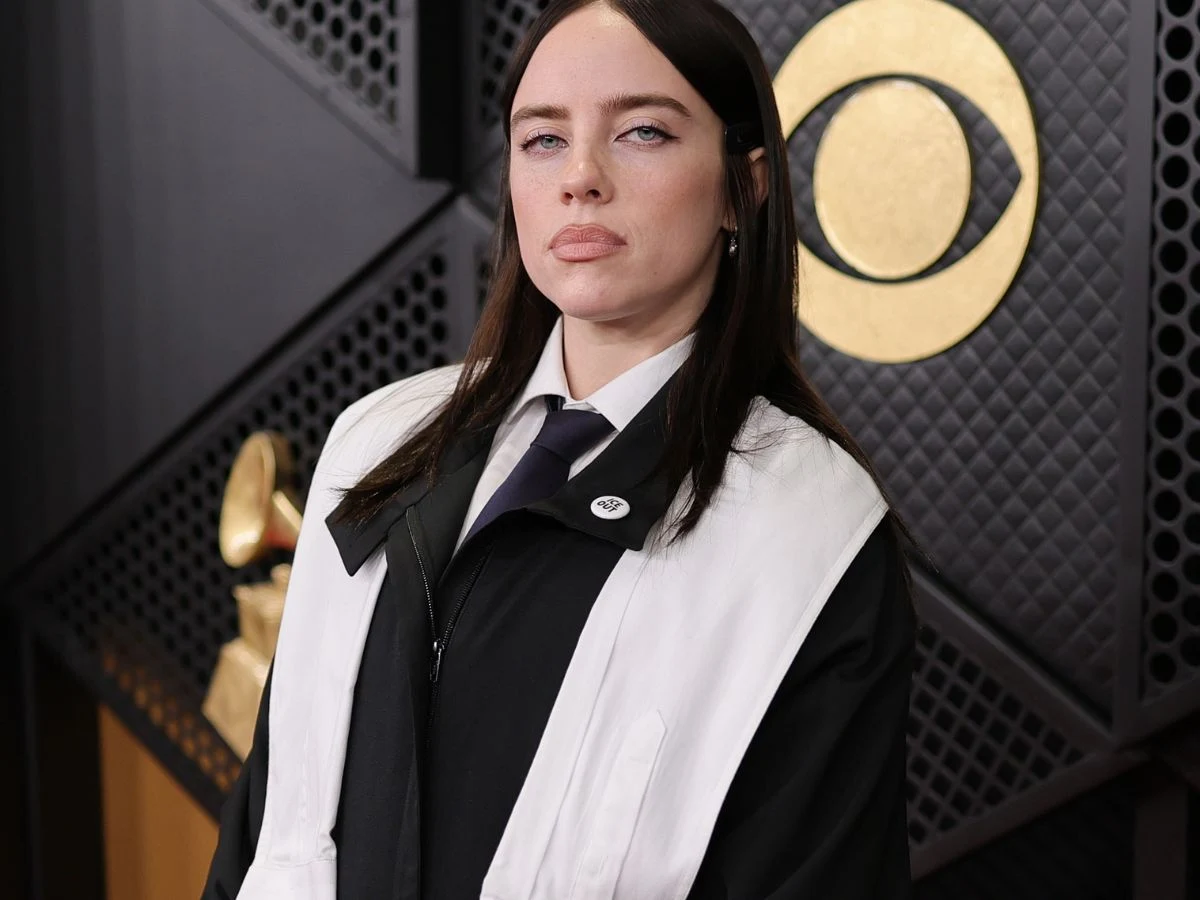 Młoda kobieta o jasnej cerze i prostych ciemnych włosach ubrana w czarno-białą stylizację z krawatem, stoi na tle ścianki z charakterystycznym logotypem CBS i złotą statuetką Grammy.