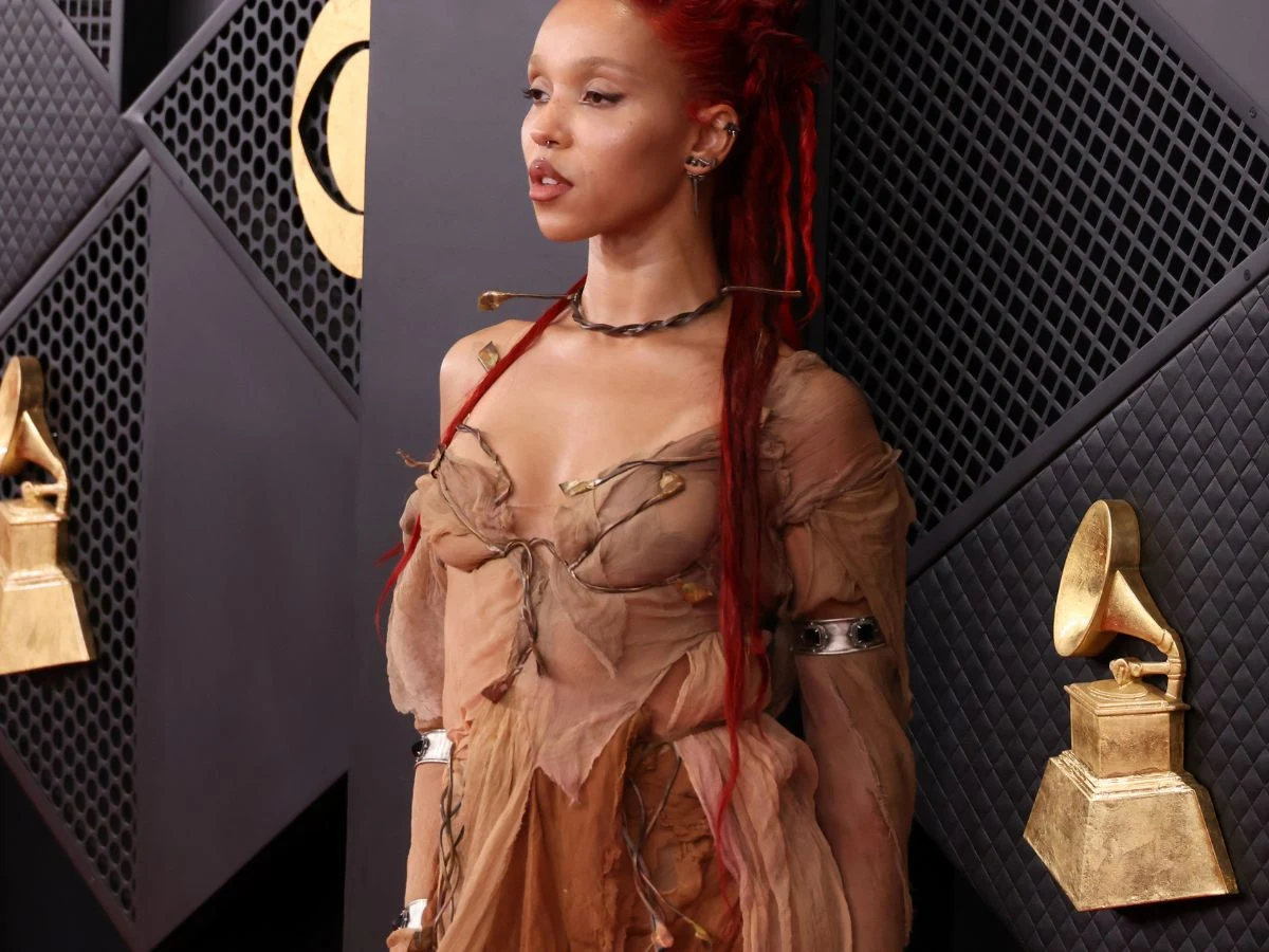 Kobieta z czerwonymi dredami w artystycznej, delikatnie prześwitującej sukni w kolorze beżowym, stojąca na tle ściany z motywem nagród Grammy, po obu stronach widoczne są statuetki Grammy.