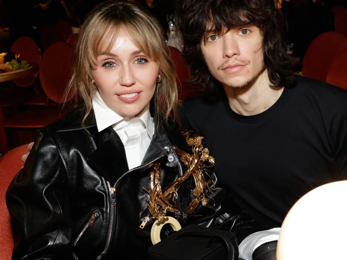 Dwie osoby siedzą przy stole, kobieta w czarnej skórzanej kurtce z ozdobami oraz mężczyzna w czarnej koszulce, patrzą w stronę aparatu, w tle widoczne rozmyte czerwone krzesła.