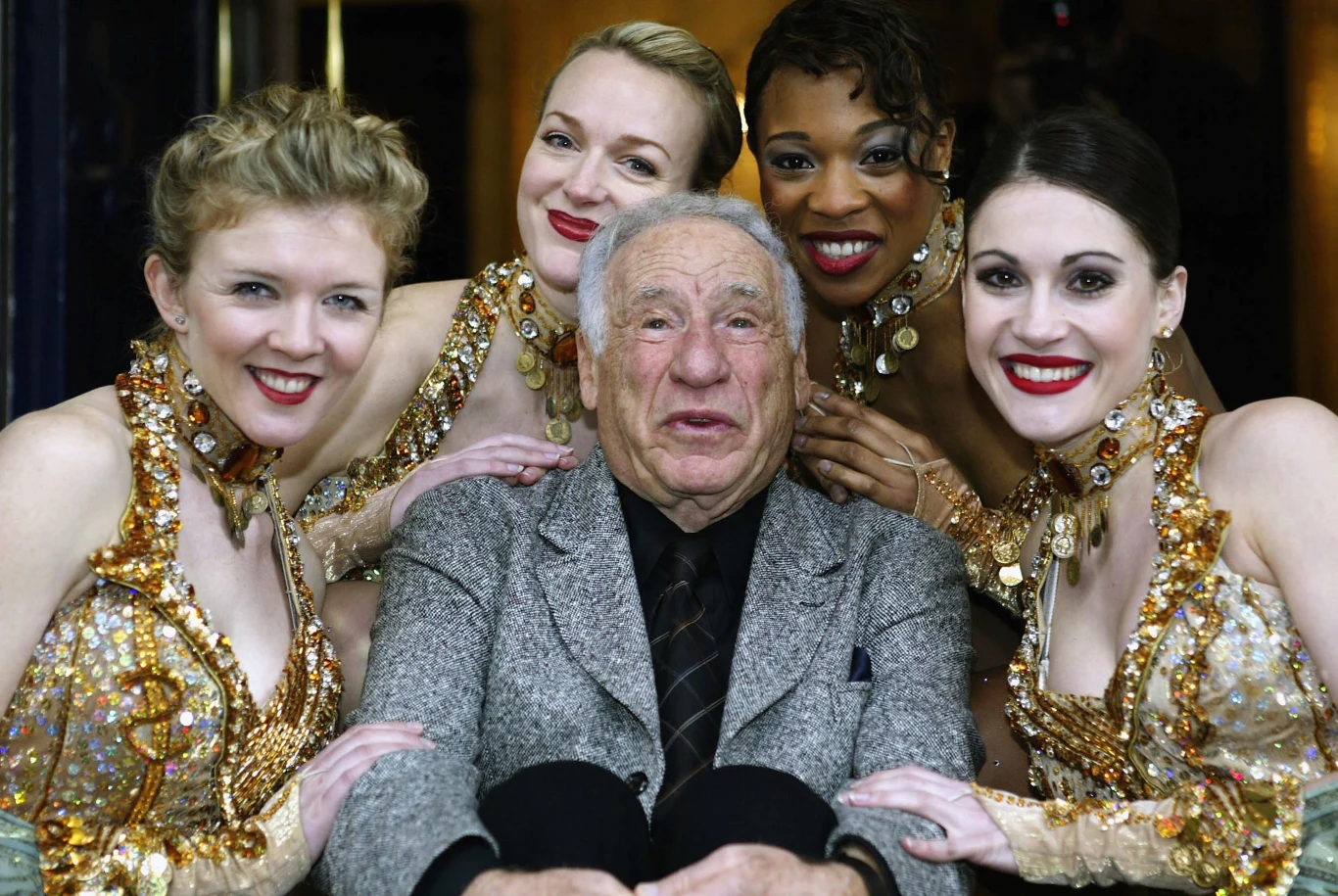 Starszy mężczyzna w centrum otoczony przez cztery kobiety w brokatowych kostiumach scenicznych. Kobiety stoją blisko niego, uśmiechają się, a ich ręce spoczywają na ramionach mężczyzny. Wszyscy prezentują radosne, teatralne nastroje.