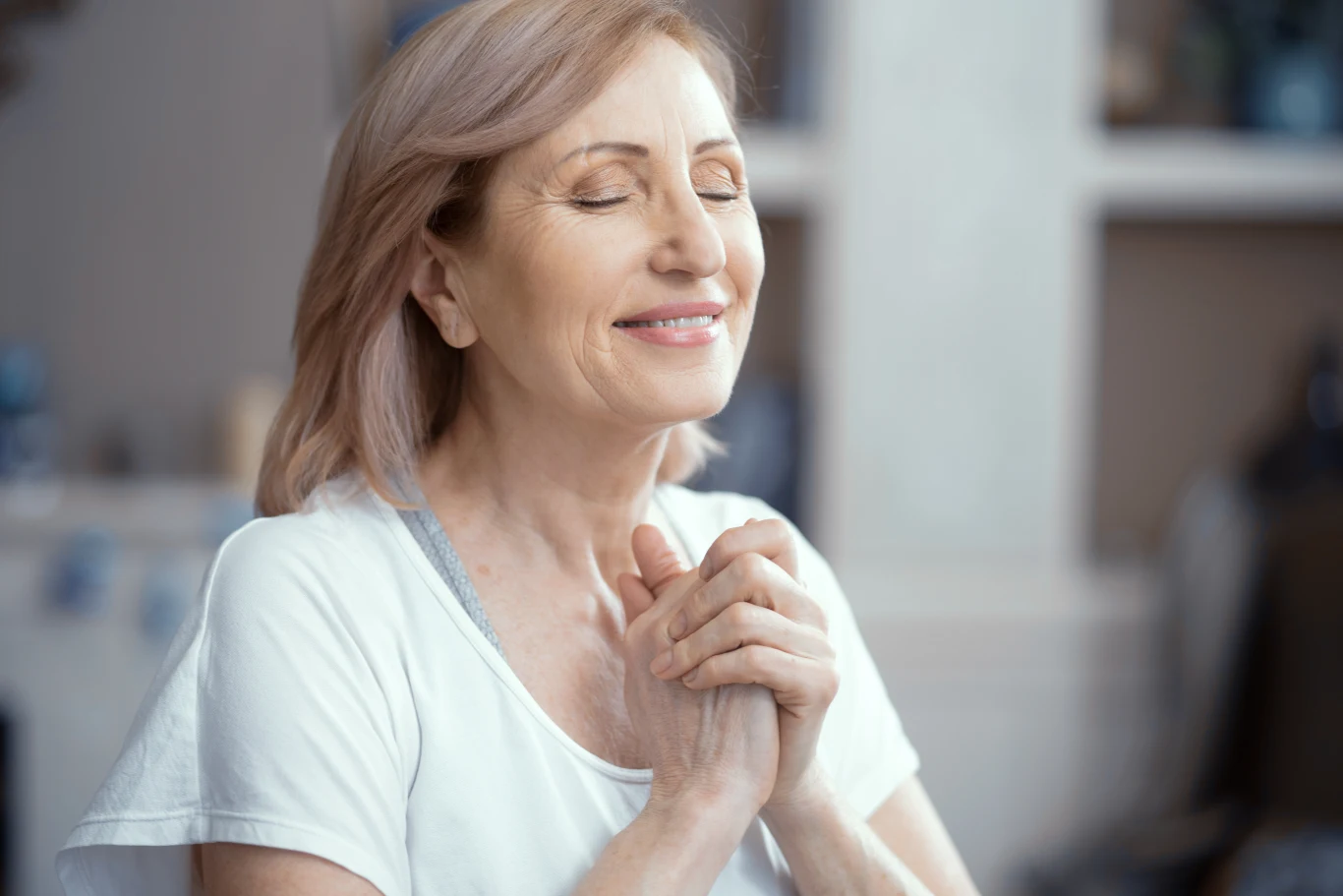 Starsza kobieta o jasnych włosach z zamkniętymi oczami i uśmiechem na twarzy, trzyma dłońmi ręce złożone w geście modlitwy lub wdzięczności. Jej postawa wyraża spokój i pozytywne emocje.
