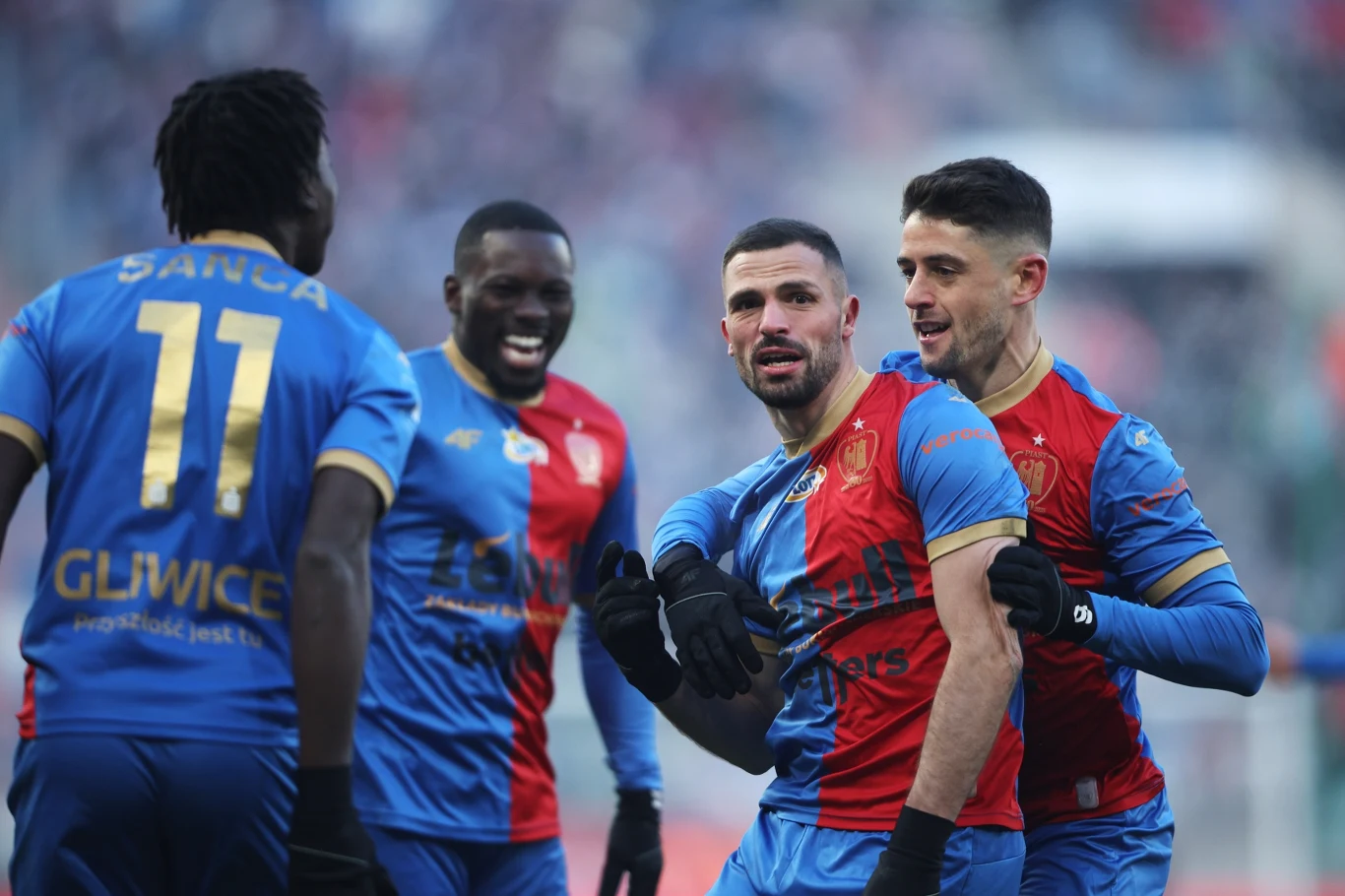 Czterech piłkarzy w niebiesko-czerwonych strojach drużyny Piast Gliwice świętuje sukces na murawie, widoczne są emocje radości i koleżeństwa.