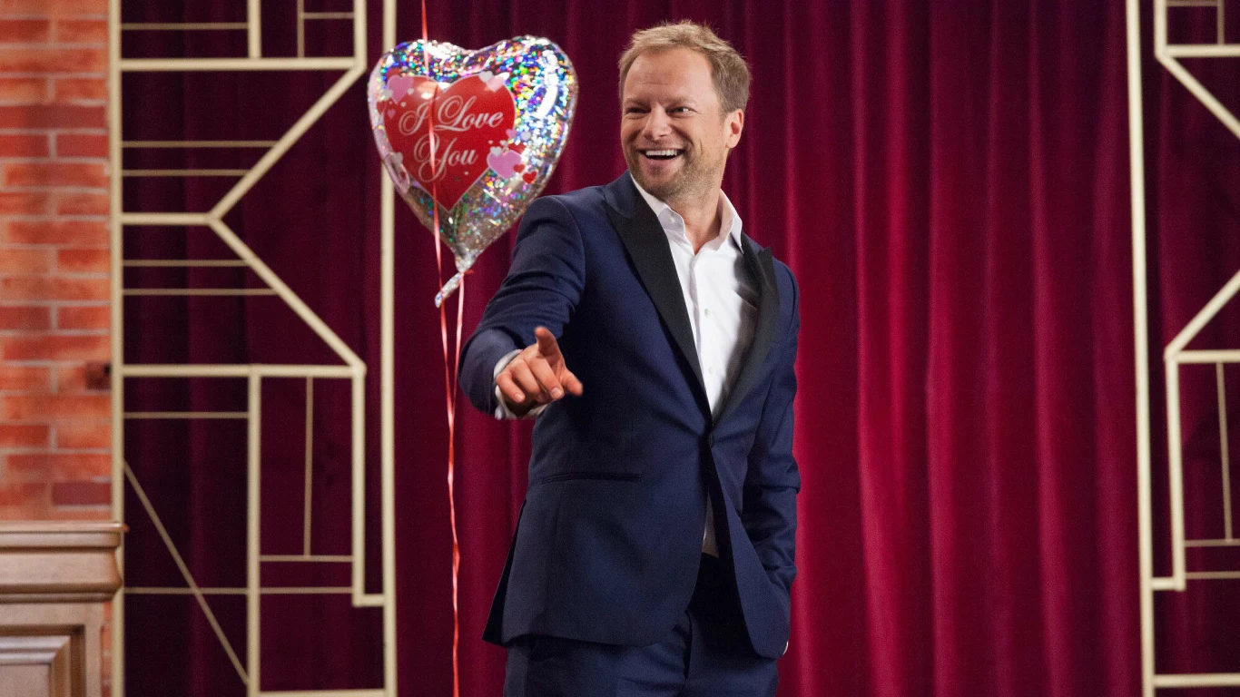 Maciej Stuhr w filmie "Planeta Singli" Mężczyzna w eleganckim granatowym garniturze uśmiecha się i wskazuje ręką, stojąc na tle czerwonej kurtyny oraz balonu w kształcie serca z napisem 'I love you'.