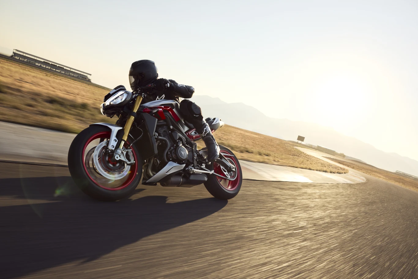 Naked o charakterze rasowej torówki. Triumph Street Tripe 765 RX ma wszystko czego potrzeba do jazdy na torze. Motocyklista w pełnym rynsztunku porusza się sportowym motocyklem po asfaltowej trasie, przy słonecznej pogodzie, z otwartą przestrzenią w tle.