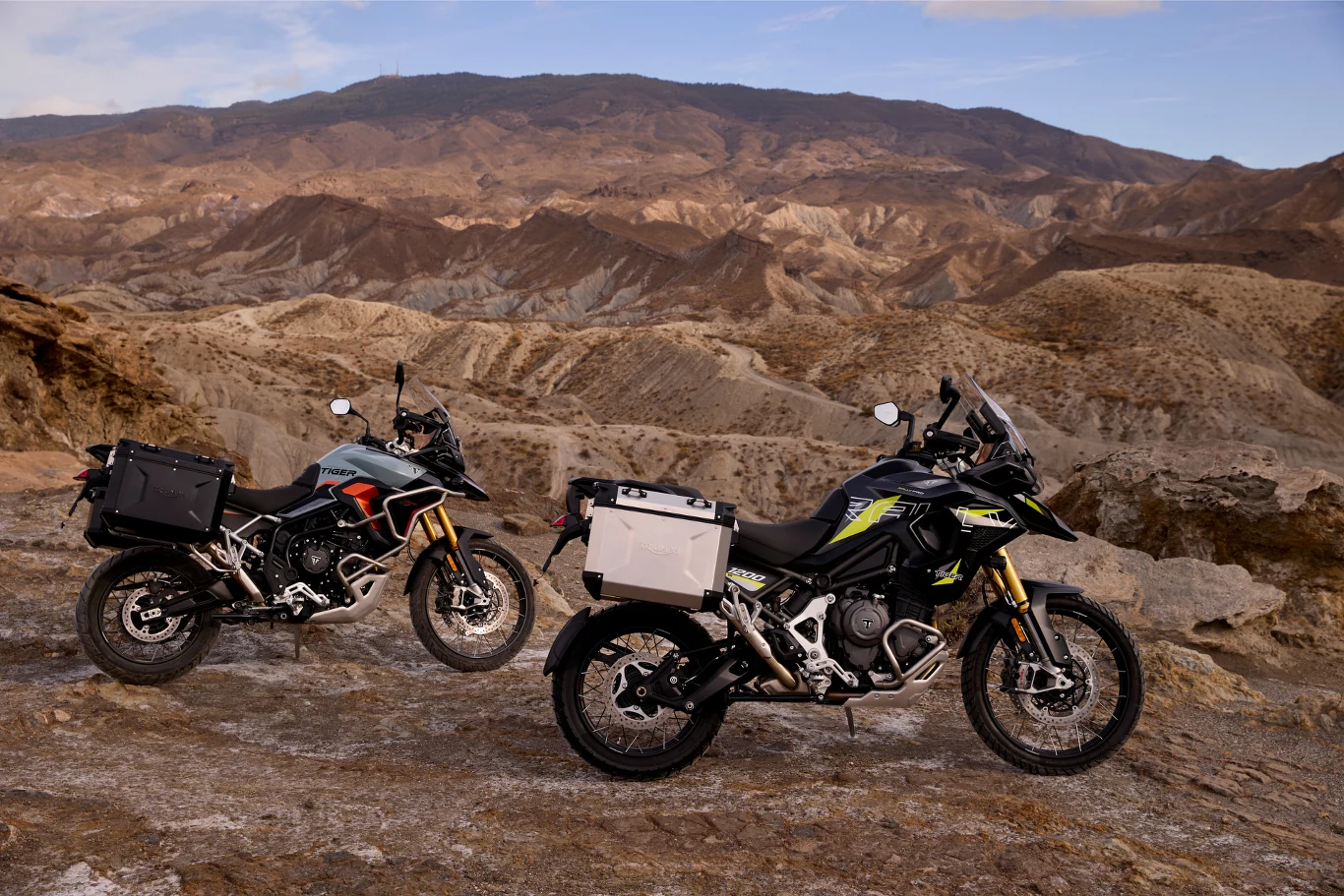 Tiger 900 i 1200 dostępny będzie w wersji Alpine i Desert. Dwa motocykle terenowe ustawione na skalistym podłożu z rozległymi, suchymi górami w tle, krajobraz pustynny podkreślający przygodowy charakter pojazdów.