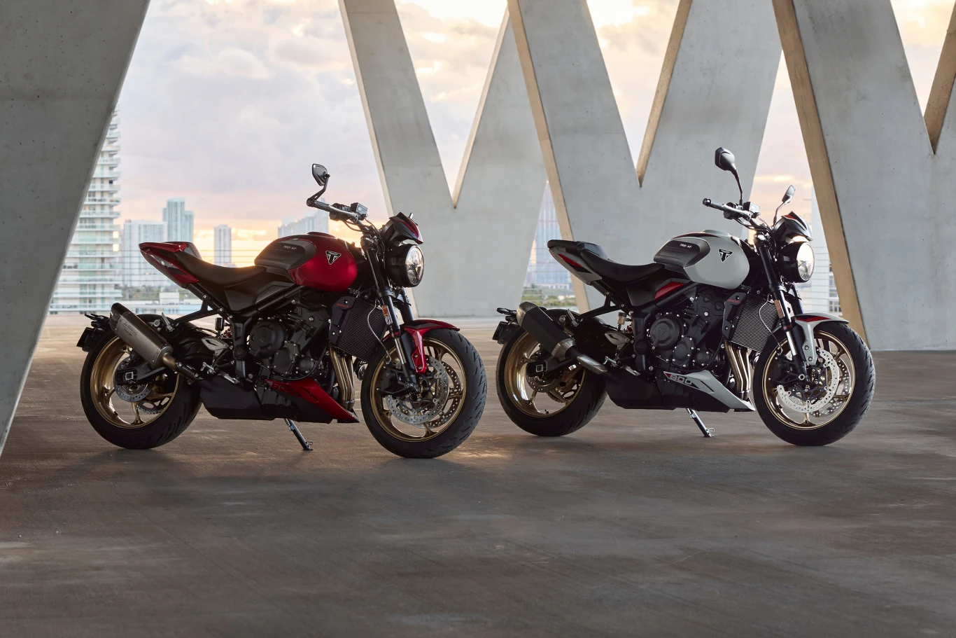 Trident 800 to nowość, na którą czekały rzesze. W ramie drzemie trzycylindrowa jednostka, której mocy nie brakuje. Dwa nowoczesne motocykle sportowe stoją na betonowej powierzchni w jasnym, przestronnym pomieszczeniu z dużymi geometrycznymi otworami w ścianie, przez które widać panoramę miasta i wysokie budynki.