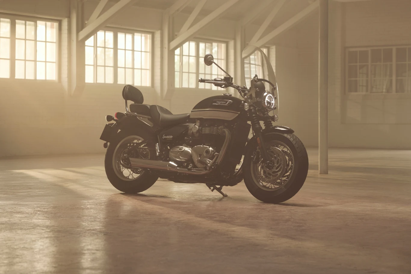 Speedmaster dostępny jest także w wersji dostosowanej do kategorii A2. Czarny motocykl o klasycznym designie stoi na środku przestronnego, jasnego pomieszczenia z dużymi oknami i widoczną stalową konstrukcją stropu. Światło słoneczne wpada przez okna, tworząc miękką poświatę na podłodze wokół pojazdu.
