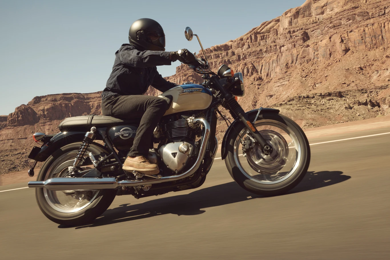 T120 jest kwintesencją rodziny Bonneville. Motocyklista w czarnym kasku i ciemnej odzieży jedzie klasycznym motocyklem po pustynnej drodze, w tle majestatyczne skały i niebieskie niebo.