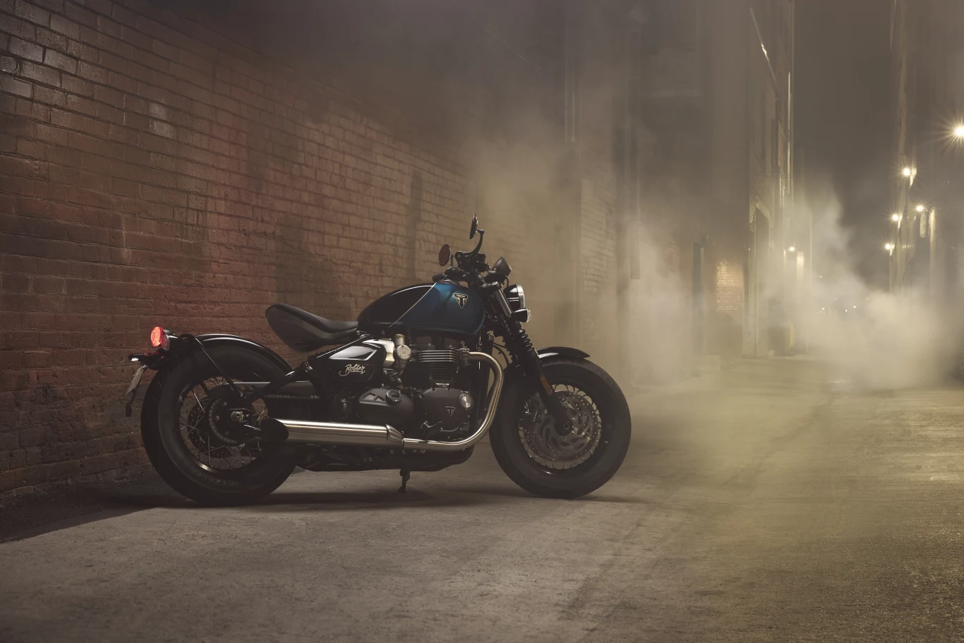 Bobber przyciąga wzrok nie tylko swoją linią, ale przede wszystkim siedziskiem. Motocykl zaparkowany przy ceglanym murze w wąskiej uliczce, po której unosi się mgła lub dym, a rozmieszczone wzdłuż ulicy lampy uliczne rzucają delikatne światło.