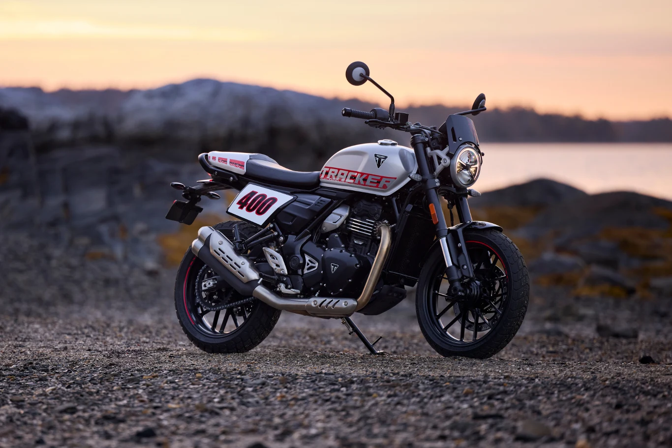 Czterysetki rosną w siłę - do gamy Triumpha dołącza Tracker 400. Motocykl typu scrambler z numerem 400 na bocznej owiewce zaparkowany na żwirowym podłożu przy zachodzie słońca, w tle rozmyte kontury wzgórz i brzegu wodnego.