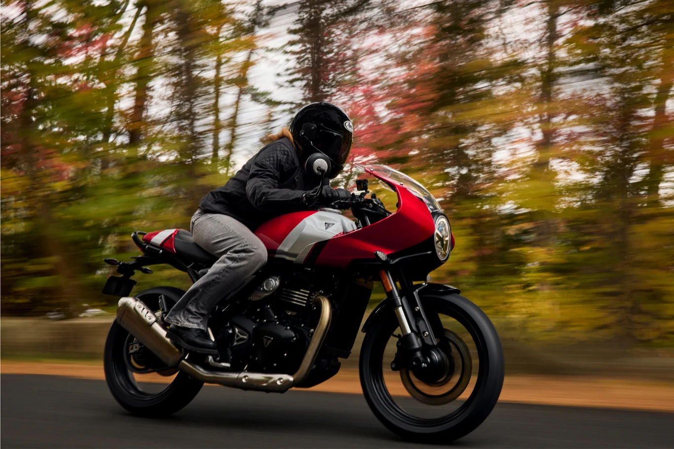 Obok tego modelu nie da się przejść obojętnie - przednia owiewka z okrągłą lampą wygląda obłędnie. Motocyklista w czarnym kasku i kurtce prowadzi czerwono-biały motocykl po asfaltowej drodze, w tle rozmyte jesienne drzewa tworzą dynamiczny efekt ruchu.
