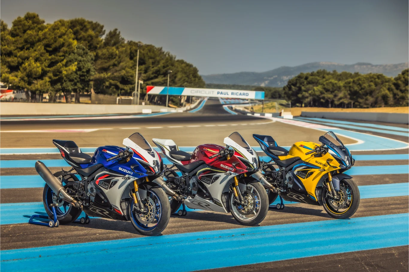 Takich głośnych powrotów chcemy! W salonach Suzuki pojawi się GSX-R1000R. Trzy sportowe motocykle w różnych kolorach – niebieskim, czerwonym i żółtym – ustawione obok siebie na torze wyścigowym Paul Ricard. W tle widoczne trybuny oraz otoczenie z drzewami i górami.