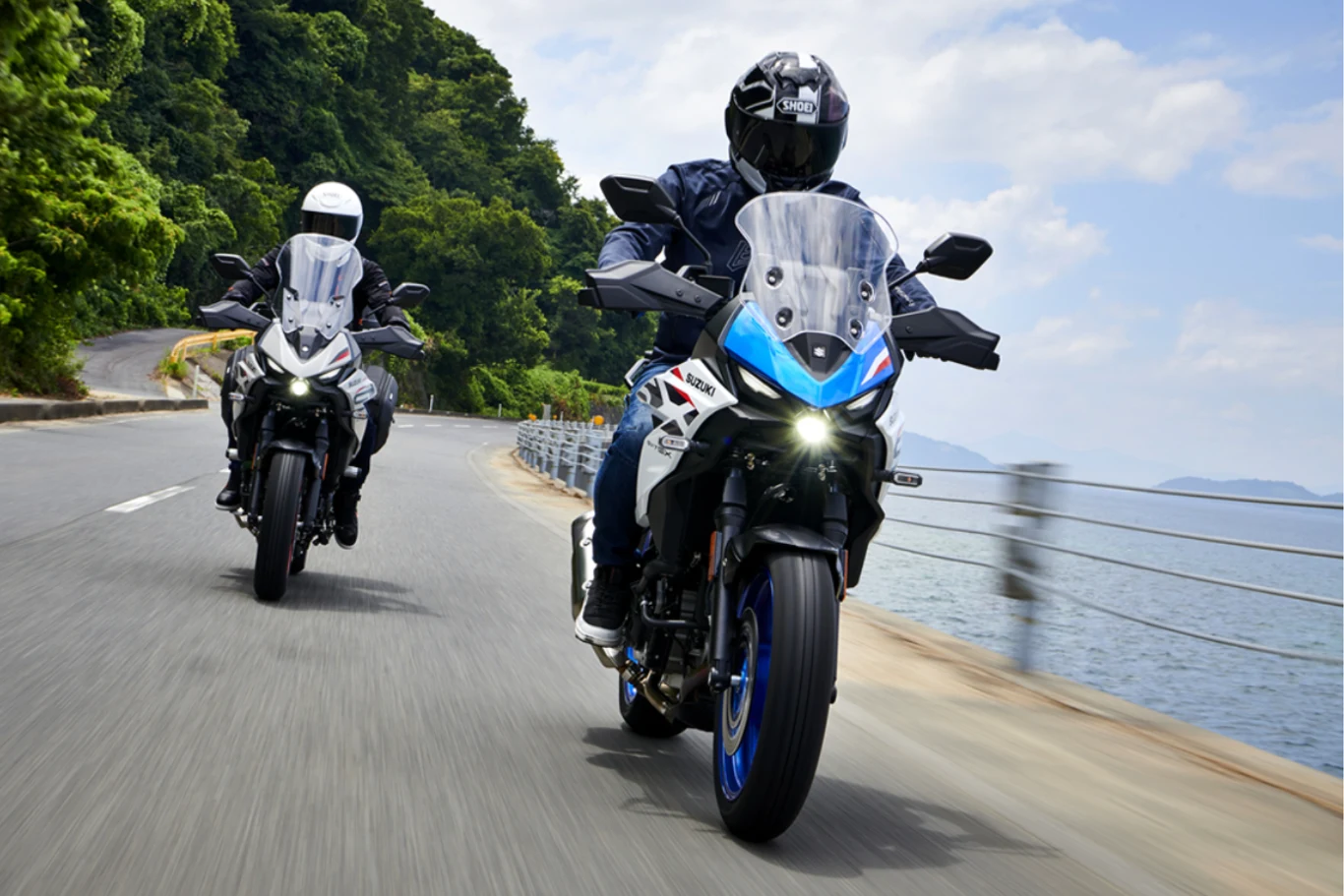 Crossover dla każdego - SV7-GX nie boi się żadnej przygody. Dwóch motocyklistów w kaskach i odzieży ochronnej jedzie dynamicznie krętą drogą wzdłuż wybrzeża, po lewej stronie gęsta zieleń, po prawej barierka i widok na wodę.