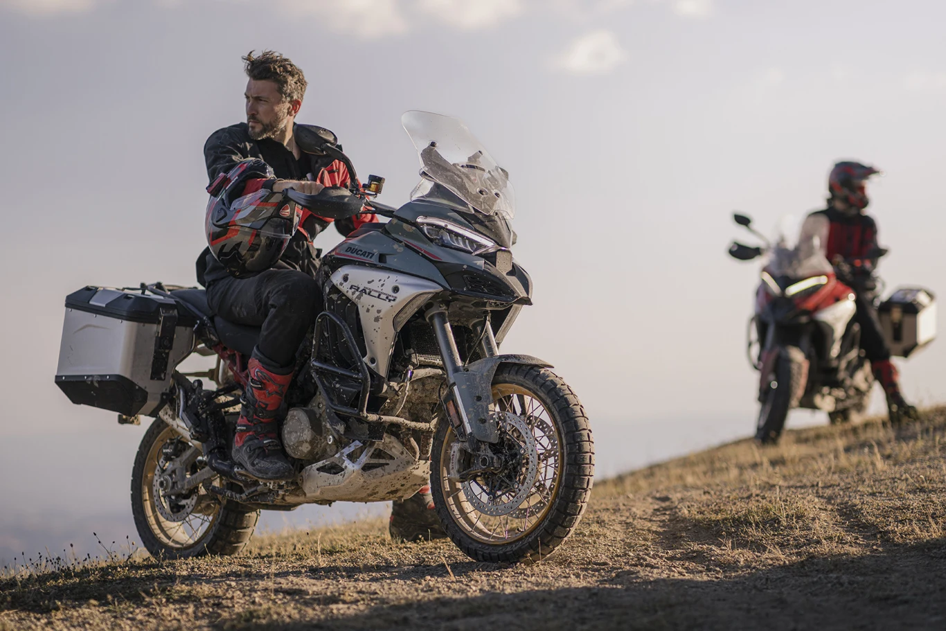 Multistrada V4 Rally zyskała jeszcze więcej wyprawowych smaczków. Dwie osoby w strojach motocyklowych jadące motocyklami terenowymi po nierównym, trawiastym terenie z lekkim nachyleniem, jedna z nich patrzy w bok, druga zatrzymała się w tle.