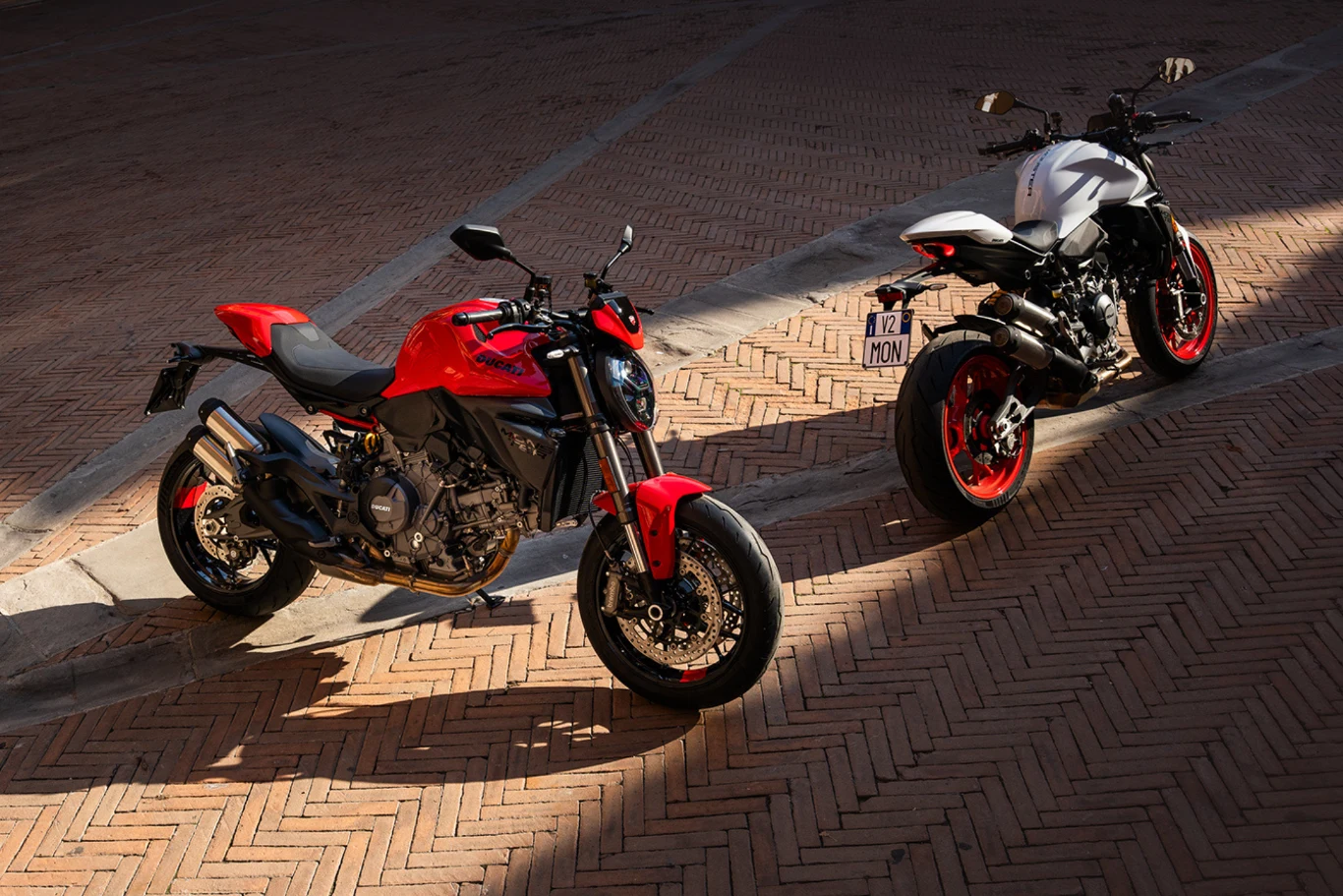 Kultowy model w nowej odsłonie - debiutuje kolejne wcielenie Monstera. Dwa nowoczesne motocykle, jeden w kolorze czerwonym, drugi biały stoją obok siebie na brukowanej powierzchni w cieniu oraz w świetle, pokazując dynamiczny design i sportowy charakter pojazdów.