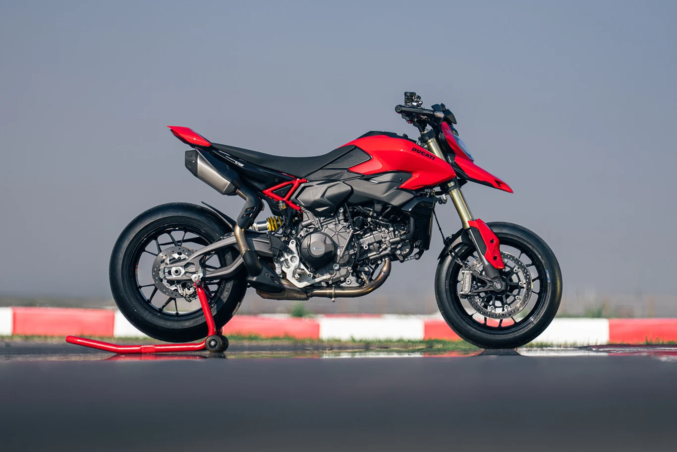 To najlżejszy i najmocniejszy Hypermotard w historii. Czerwony motocykl sportowy typu supermoto ustawiony na stojaku serwisowym na torze wyścigowym, widoczny w ujęciu bocznym na tle rozmytego nieba i barier z biało-czerwonymi pasami.